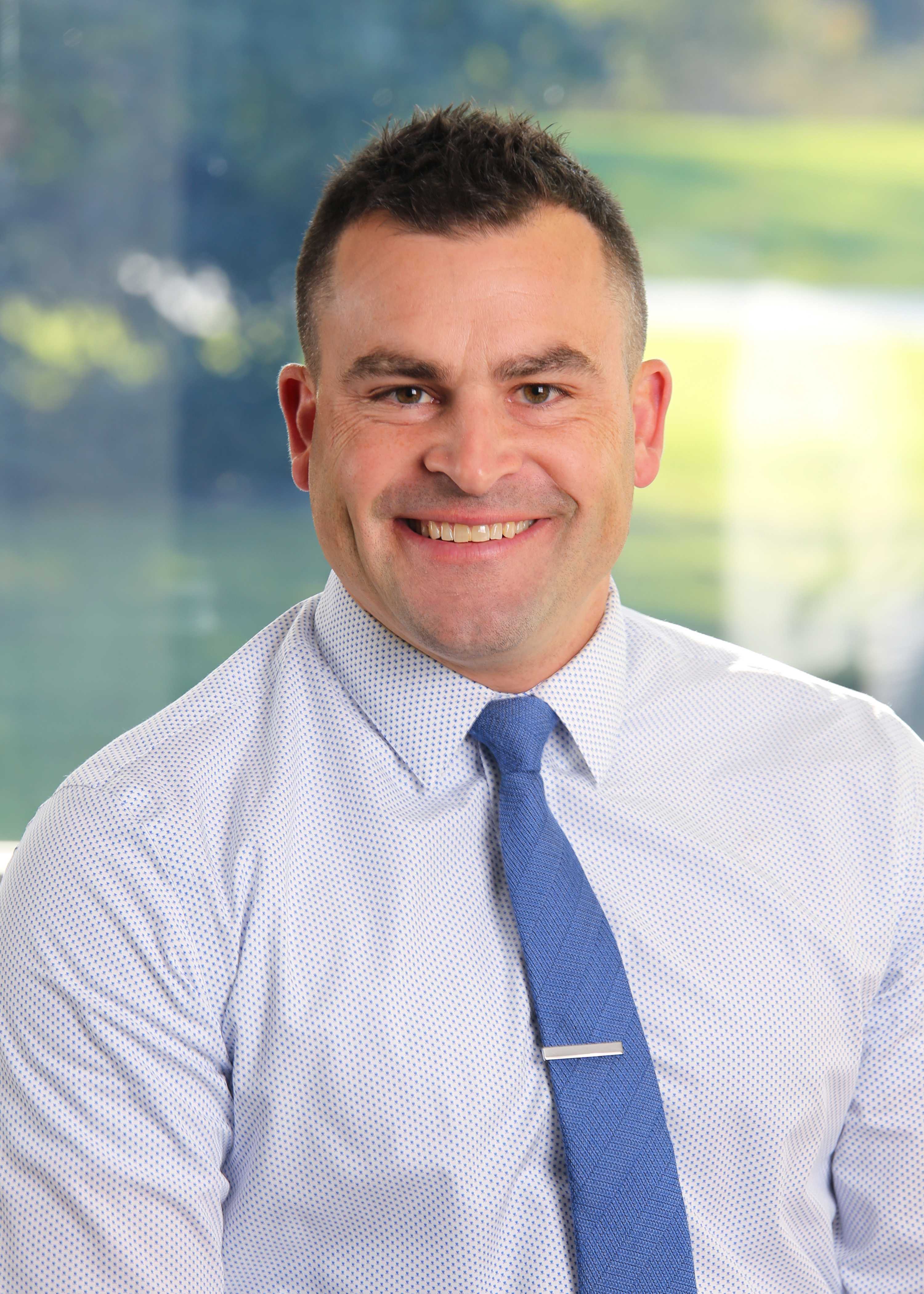 A smiling man in a light blue striped shirt and blue tie poses for a portrait.