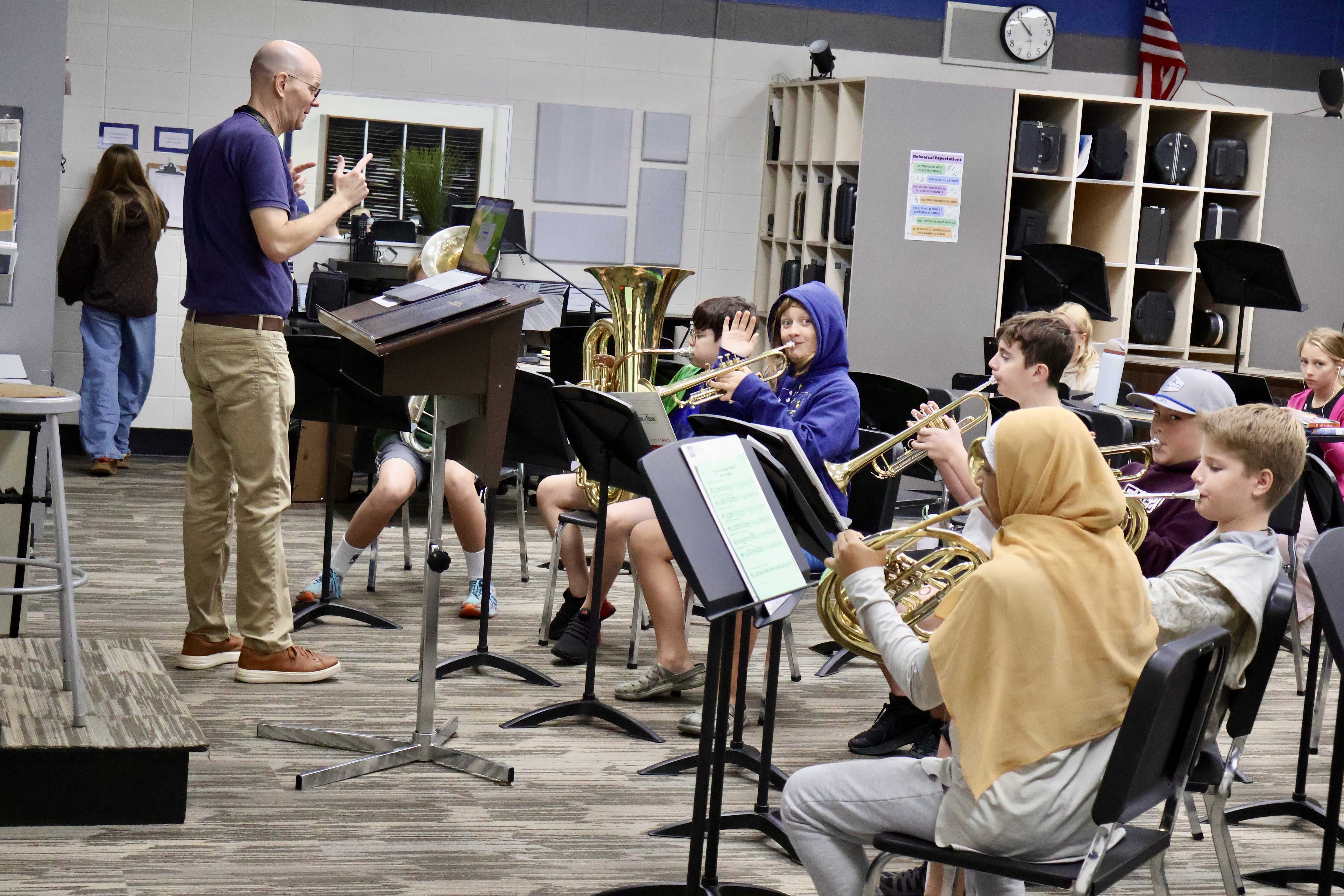 A music teacher stands at the front of a school band room, guiding a group of middle or high school students playing wind instruments.