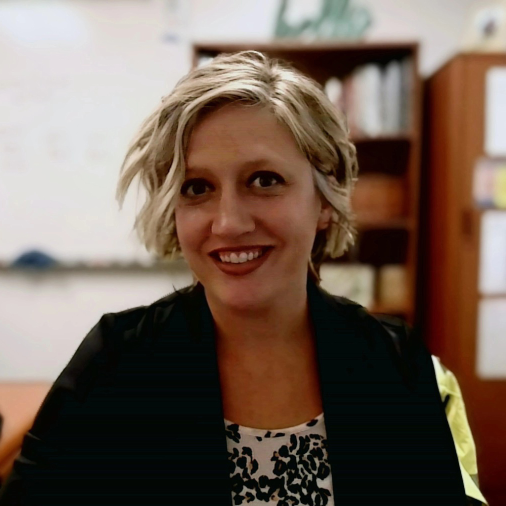 A woman with short, blonde, wavy hair smiles at the camera, wearing a black jacket over a patterned top.
