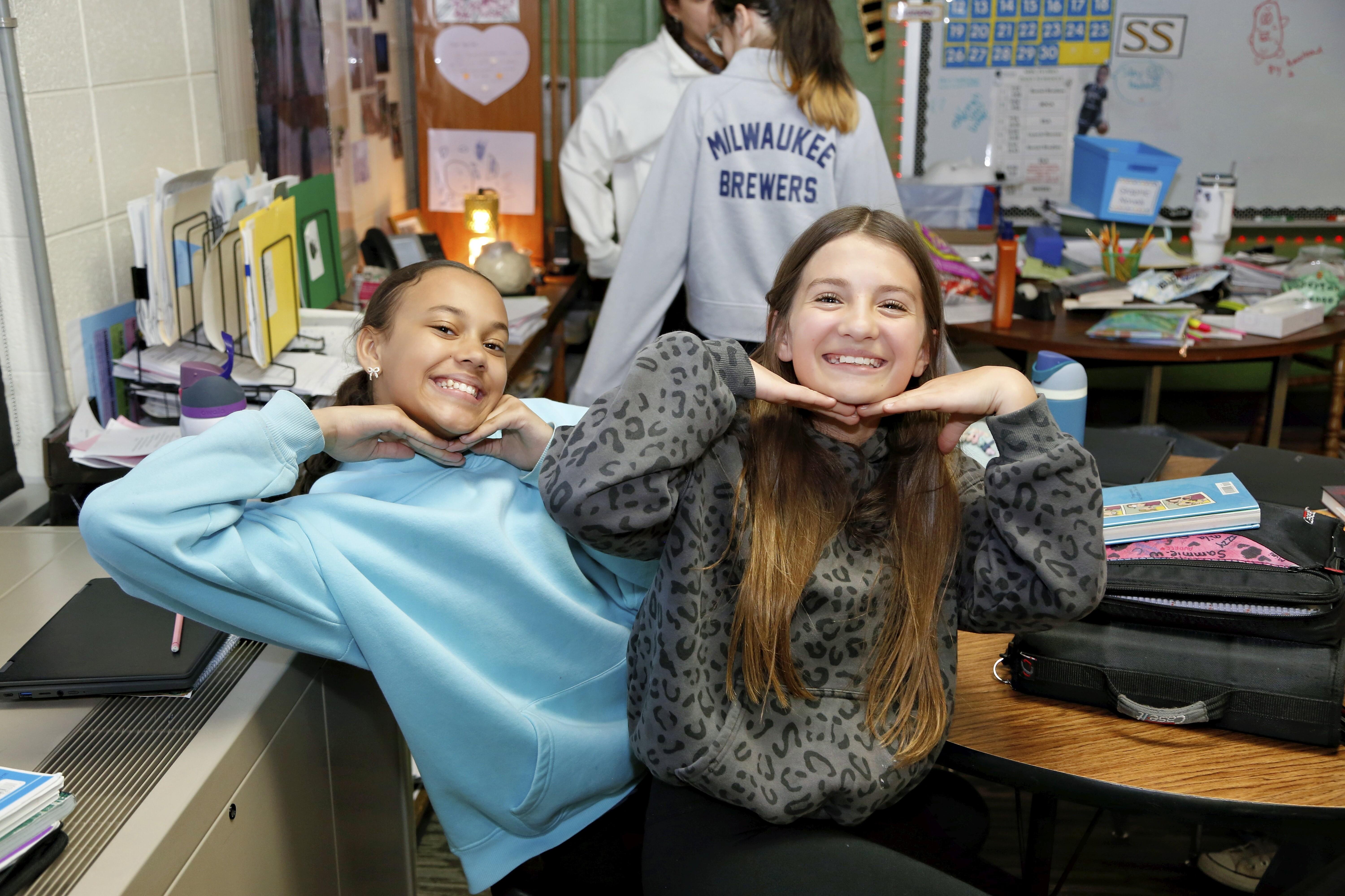 Two smiling girls in a classroom make funny faces at the camera.