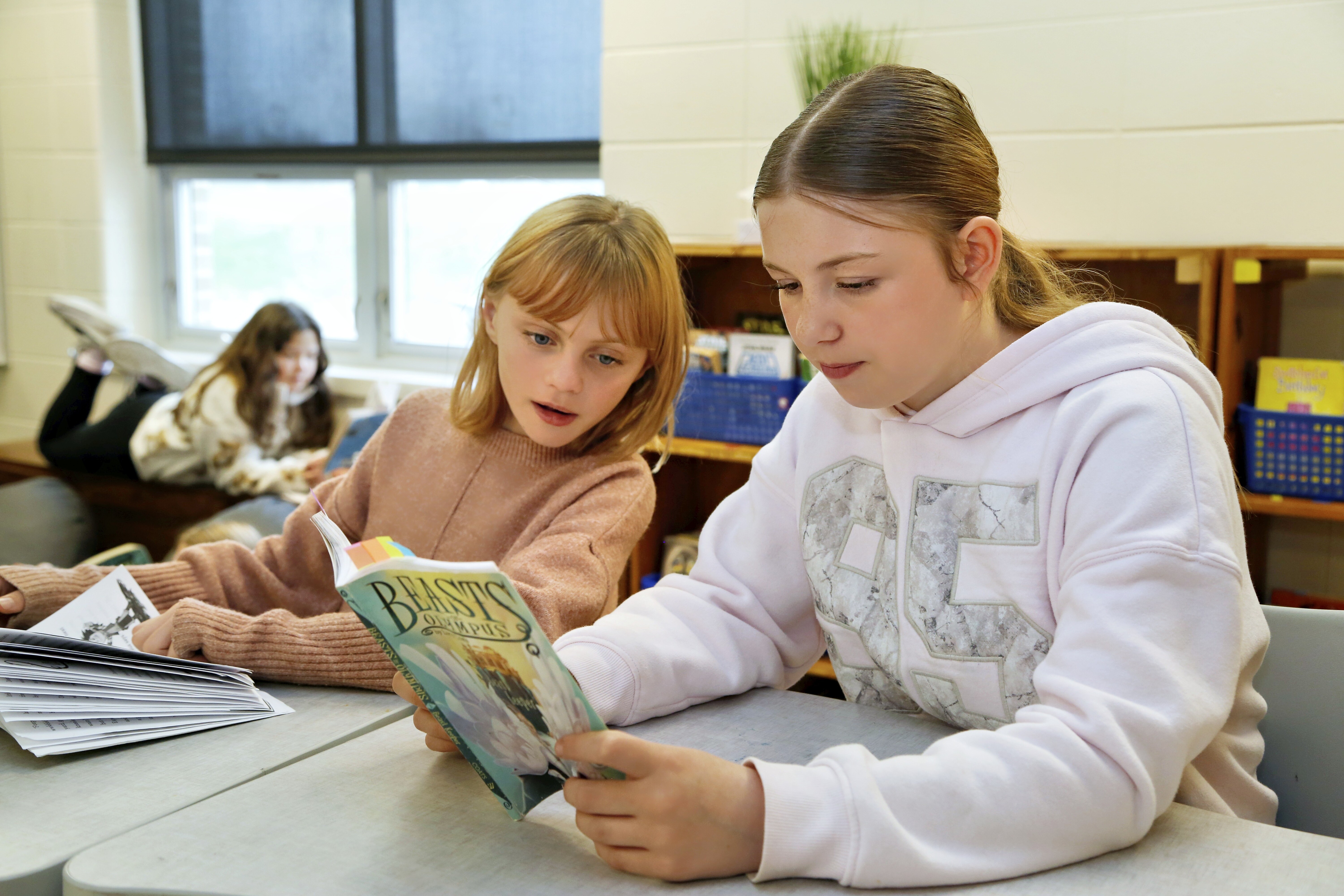 Two young girls focus on a book together at a table in a classroom.