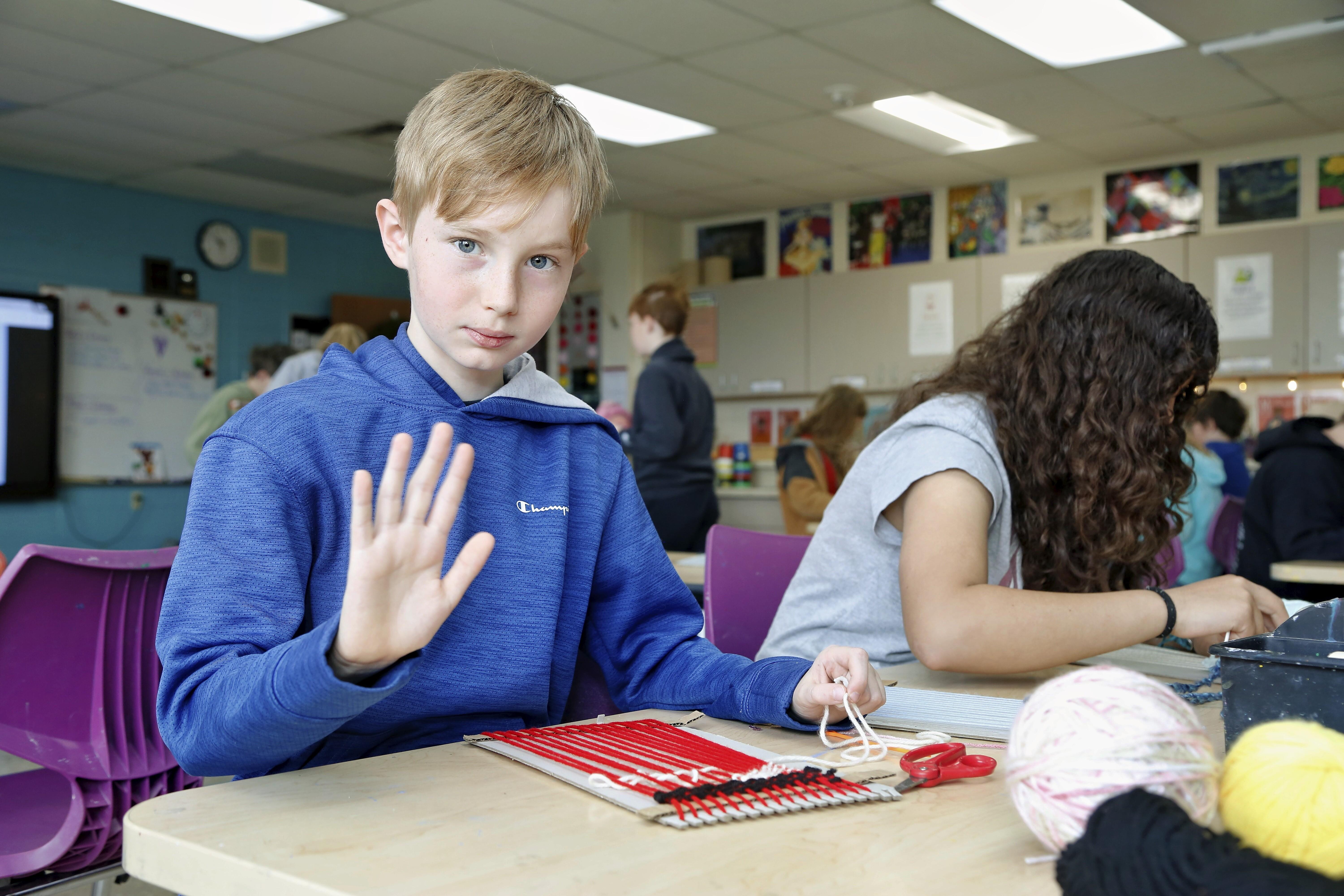 A young boy in a blue hoodie raises his hand in a classroom setting.