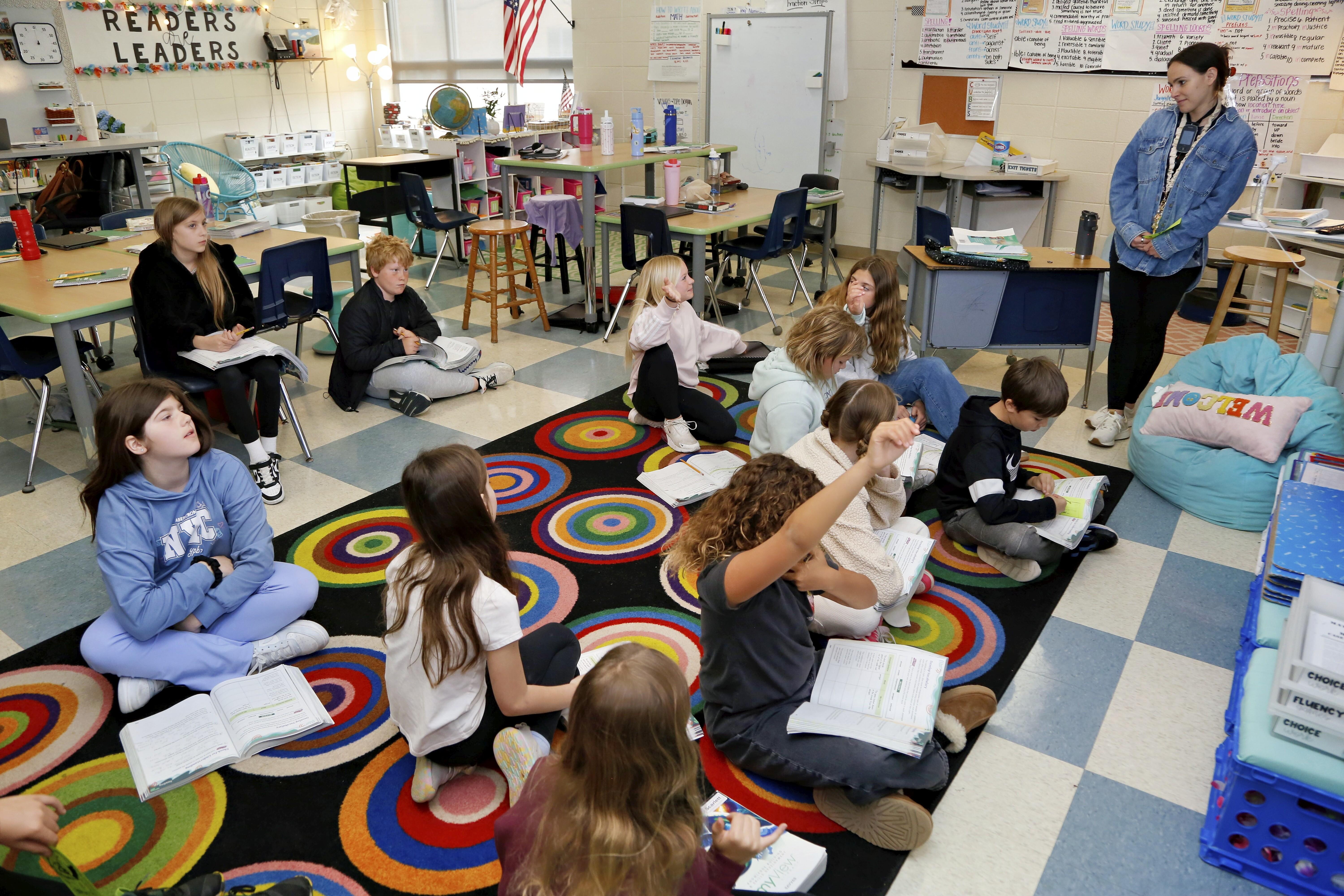Children sit on a colorful rug in a classroom, listening to a teacher.