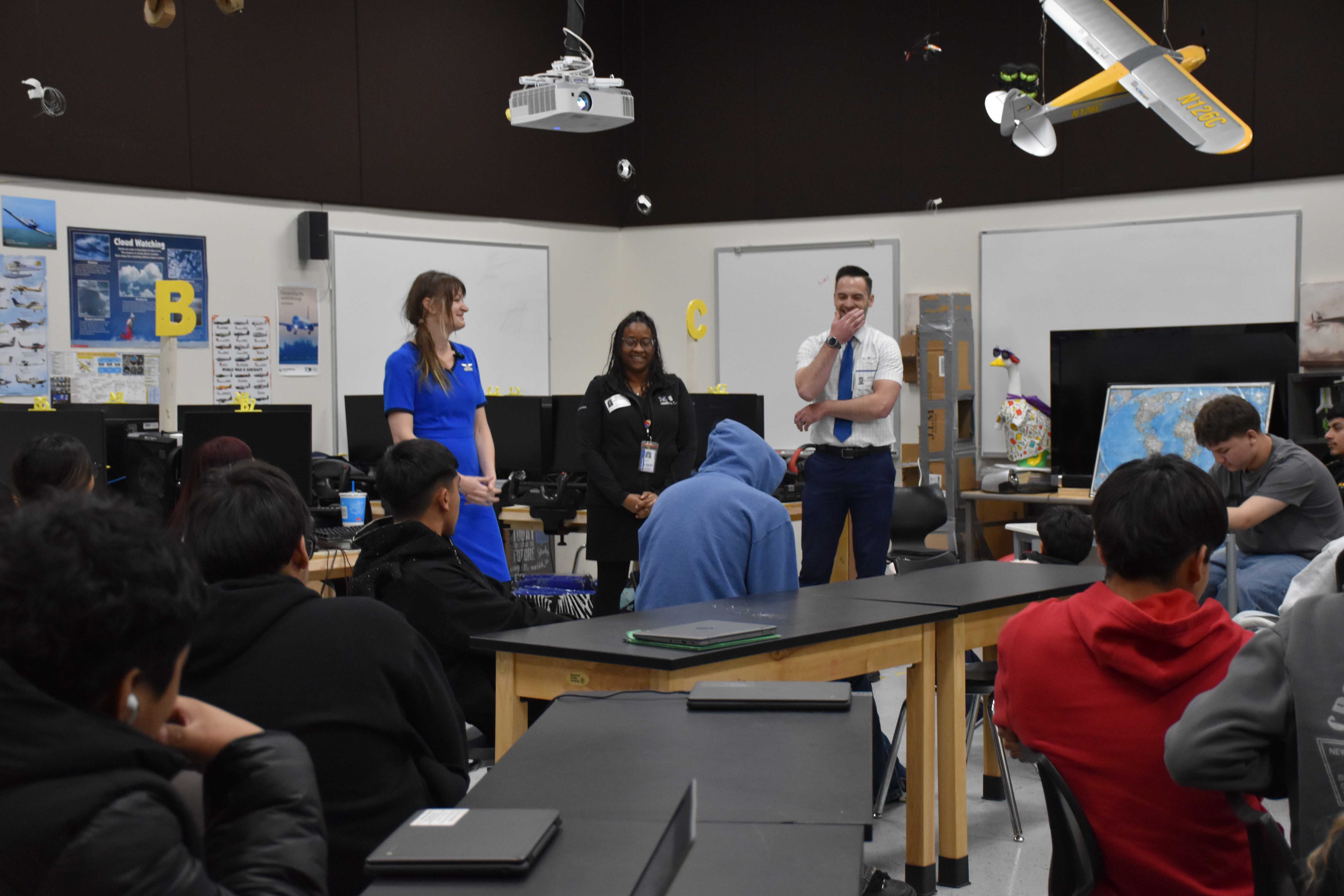 Three adults in professional attire present to a classroom of students, with a model airplane hanging overhead.