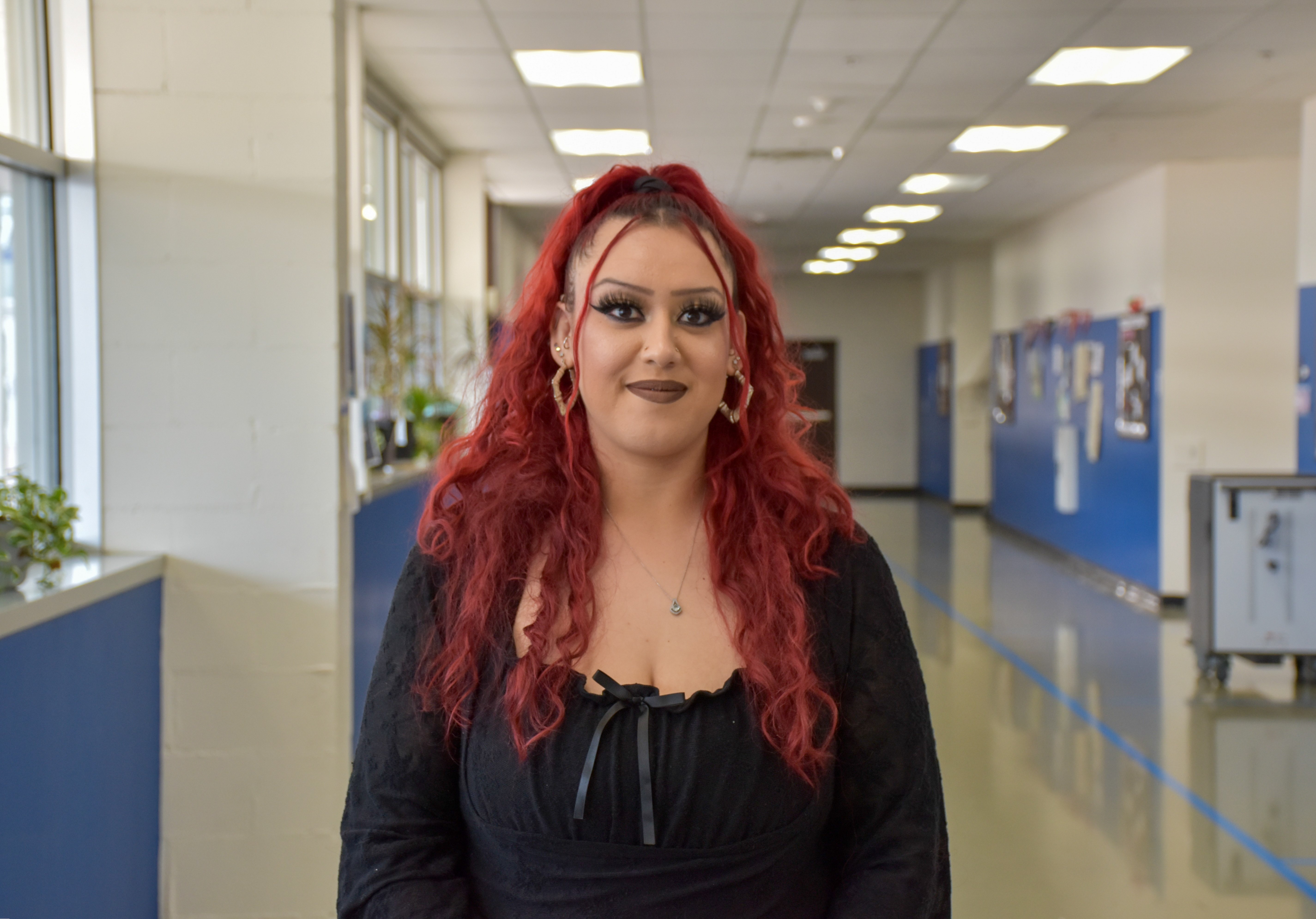 A young woman with vibrant red hair and glasses stands in a school hallway.