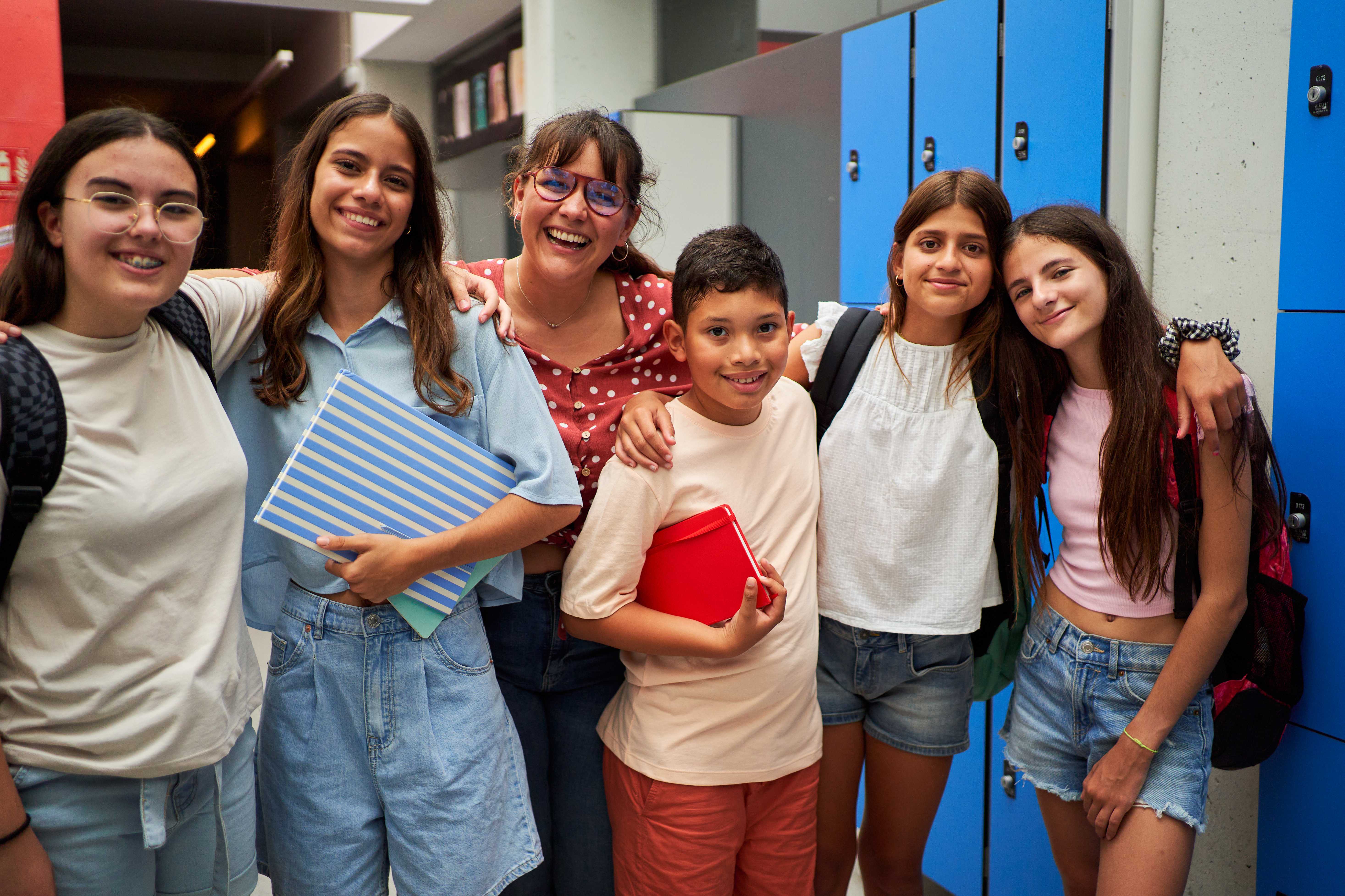 A group of smiling students and a teacher stand together in a school hallway with lockers in the background.