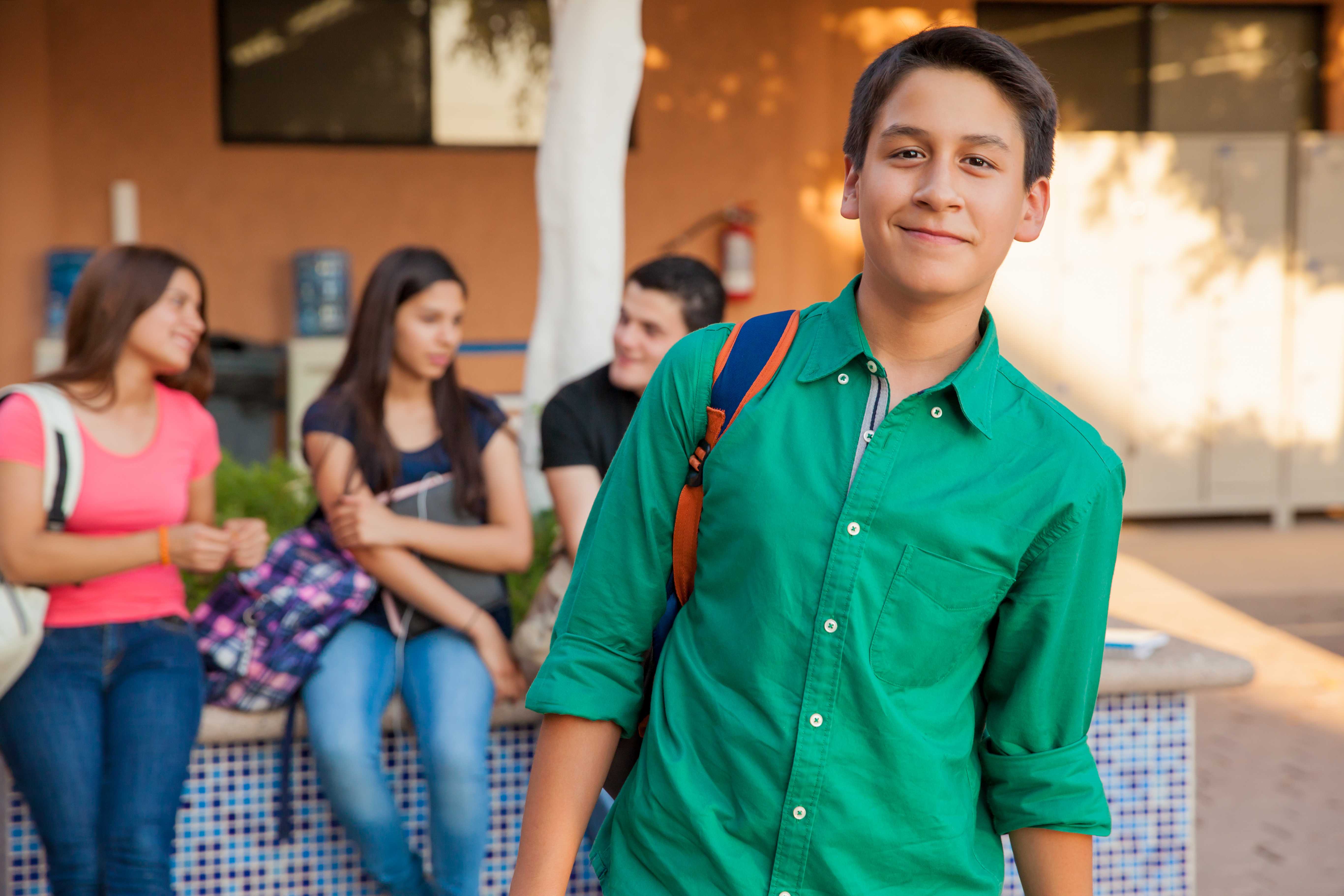 A young man in a green shirt with a backpack smiles at the camera, with other students in the background.