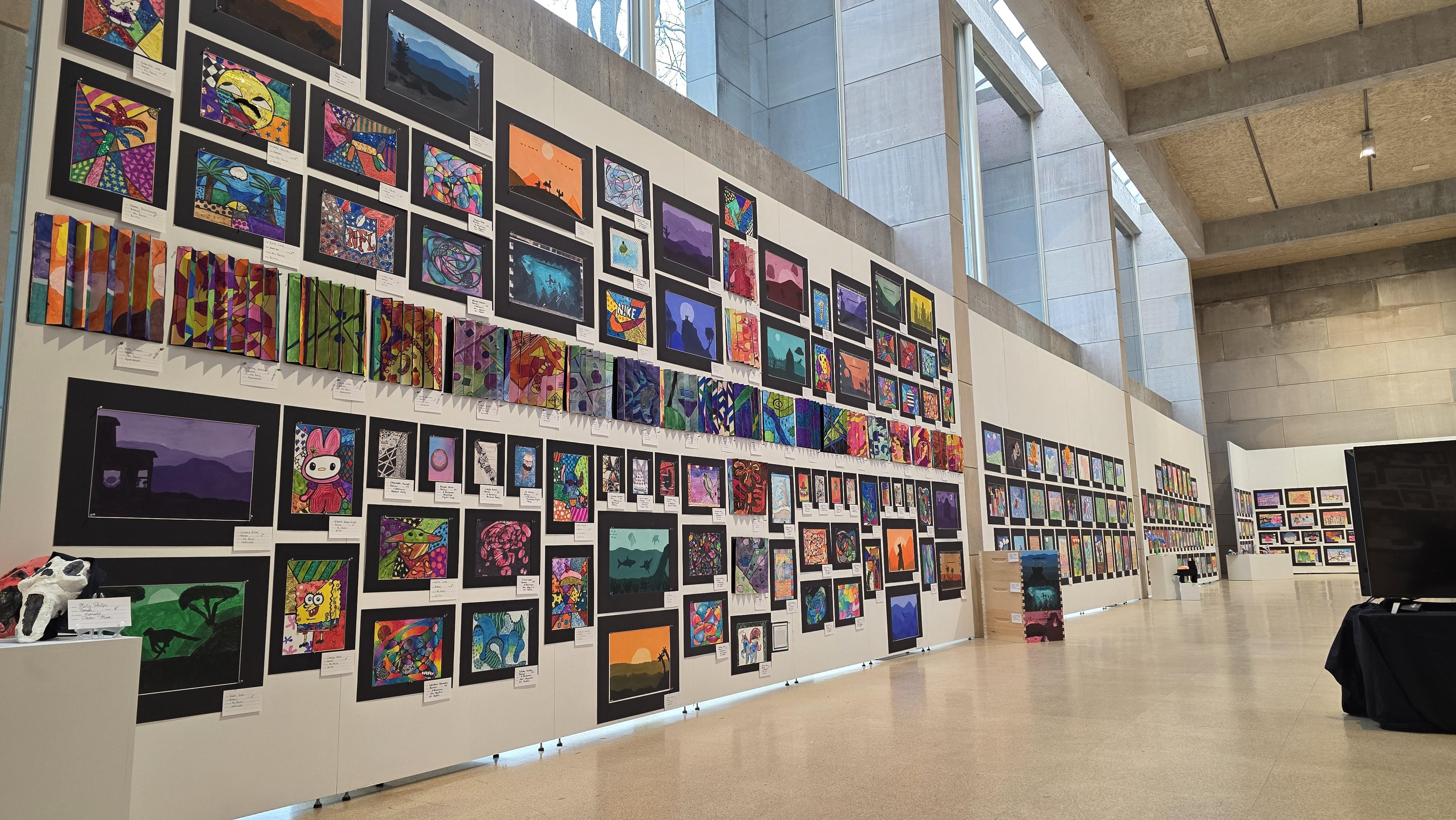 A large wall displays a grid of colorful framed artworks in a gallery setting.
