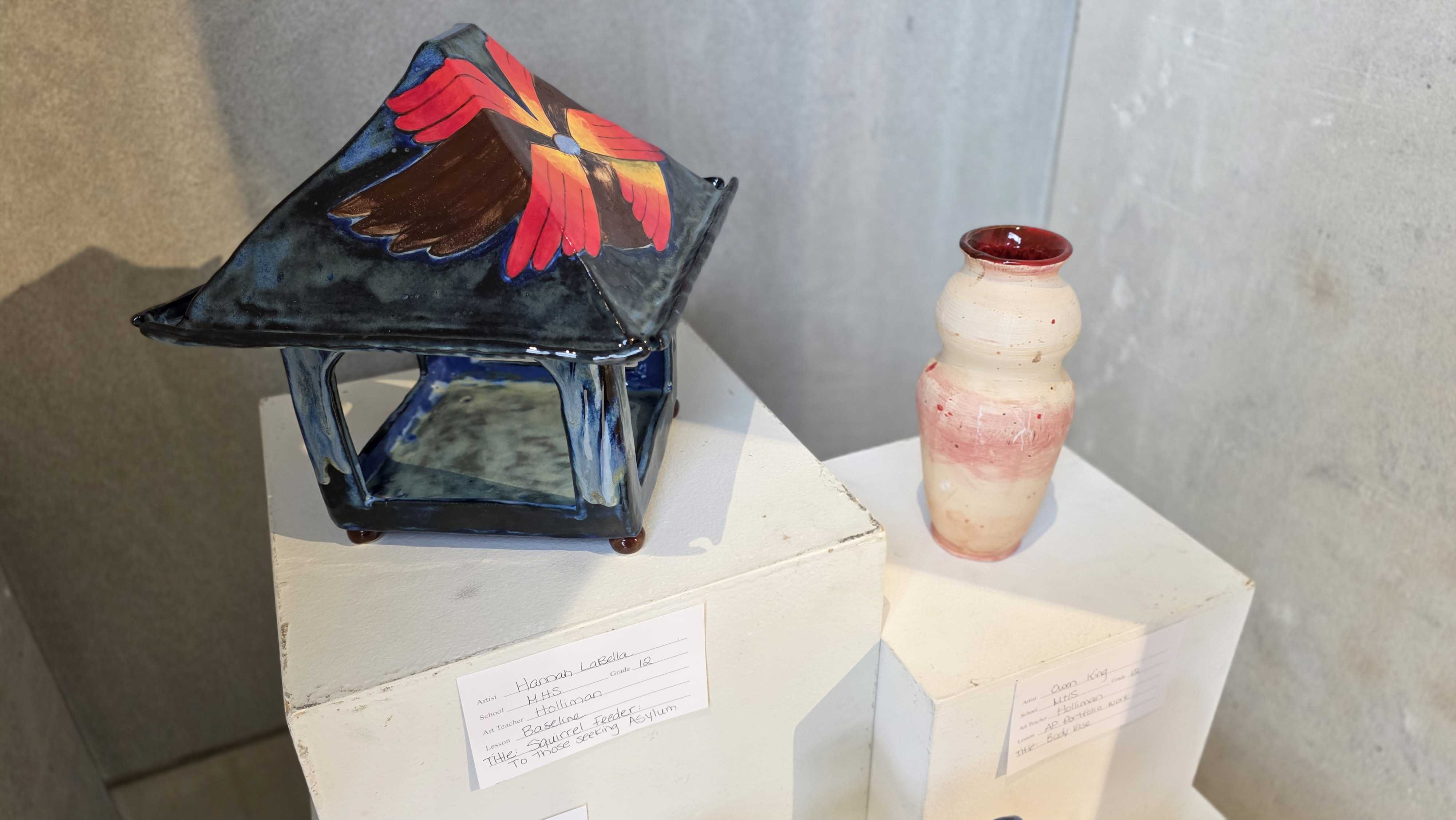 A decorative glass lantern with a peaked roof sits next to a pink and white glass vase.