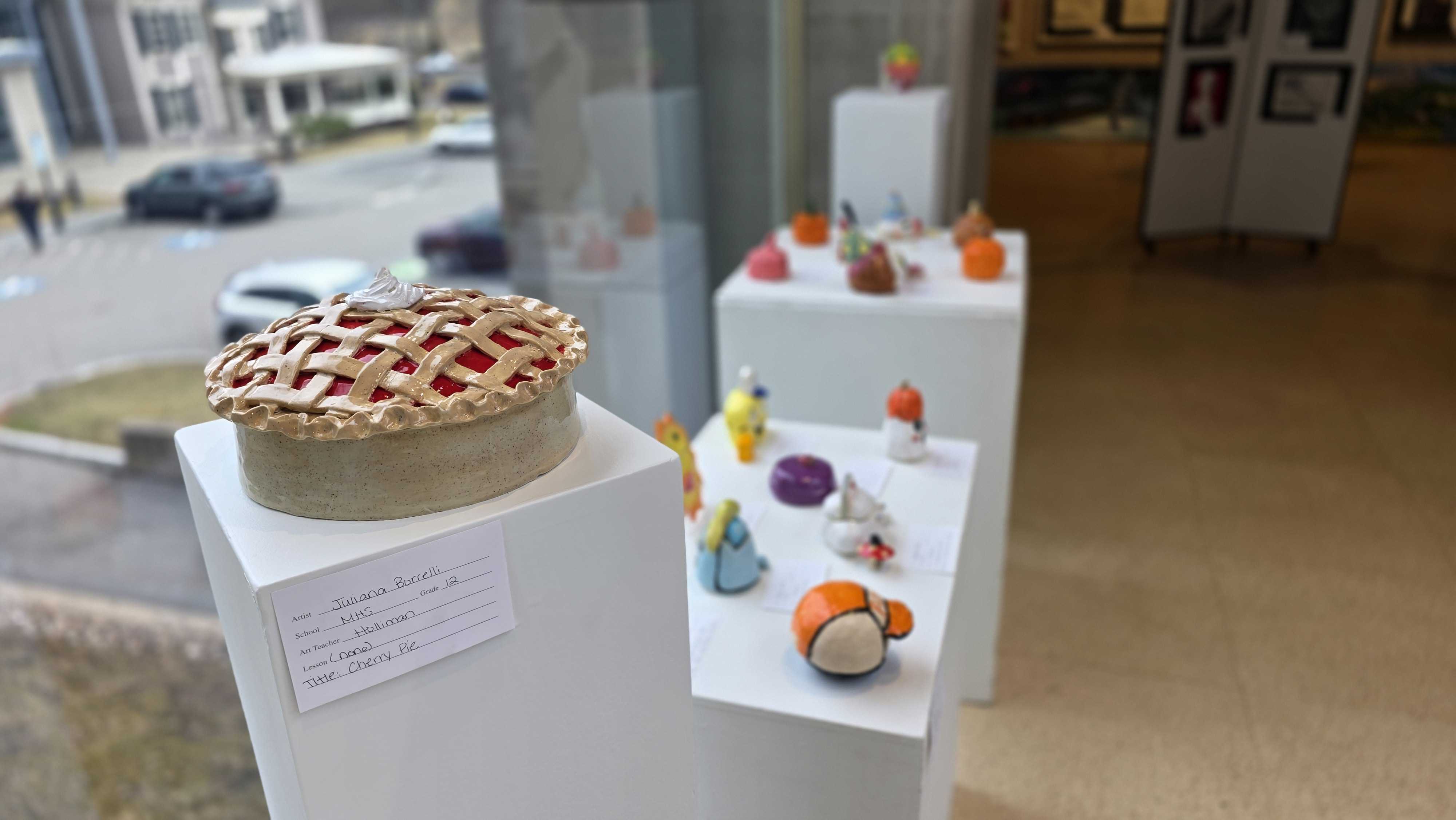 A realistic pie sculpture is displayed on a white pedestal in an art exhibition.