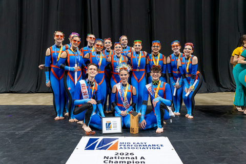 A group of performers in matching blue and orange uniforms pose for a photo with trophies.