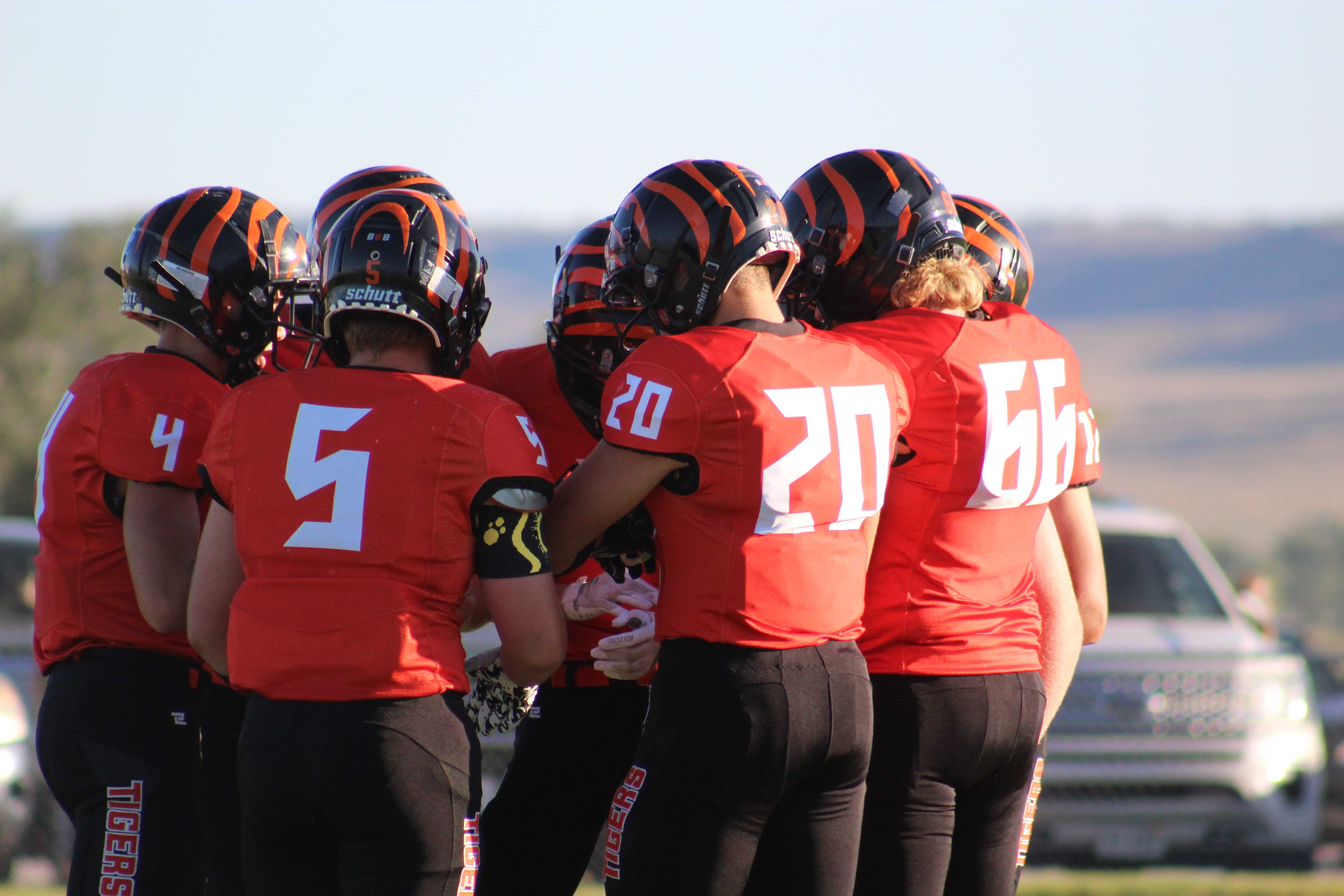 A football team wearing orange jerseys and tiger striped helmets gathers closely in a huddle on the football field.