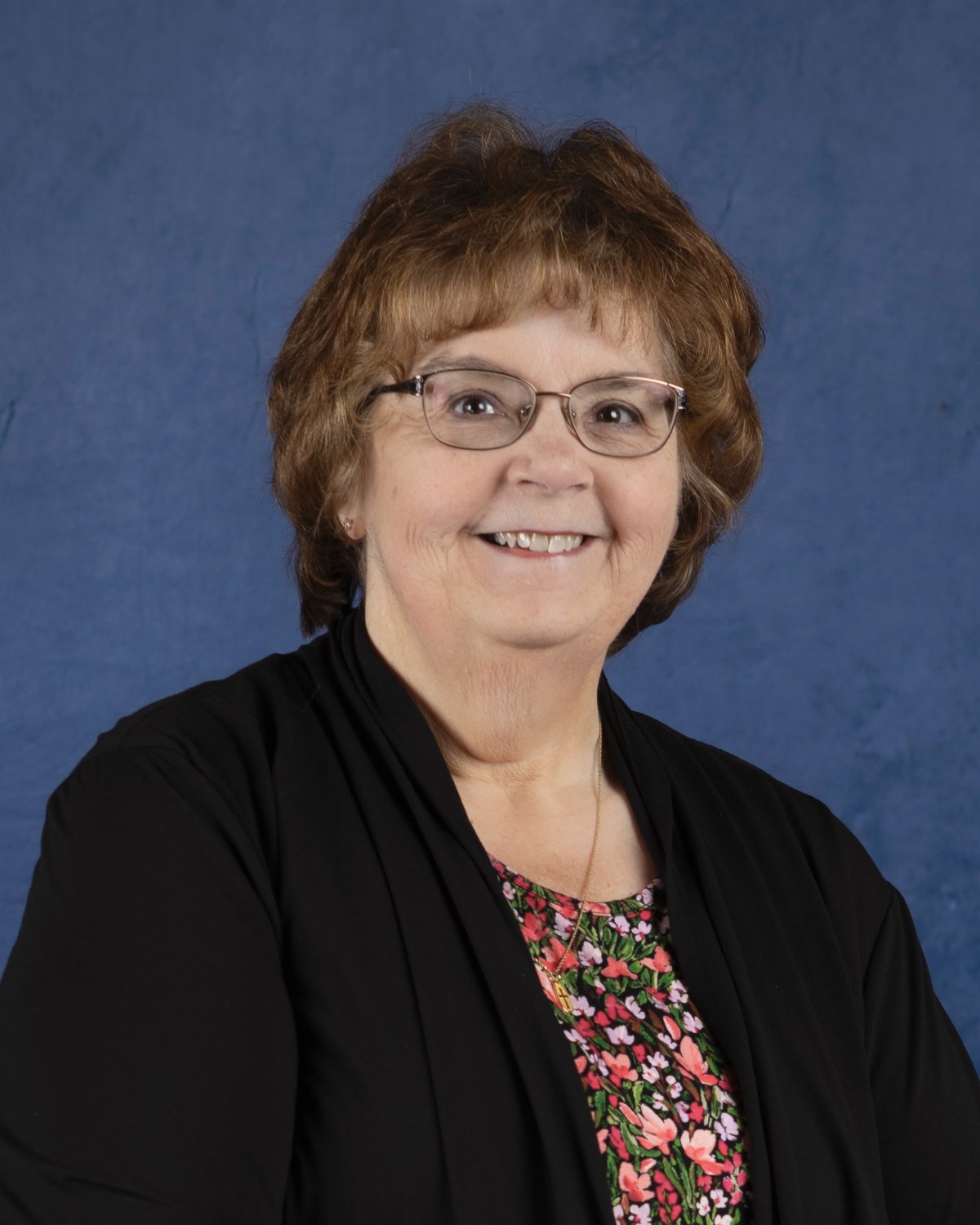 A smiling woman with brown hair and glasses poses against a blue background.