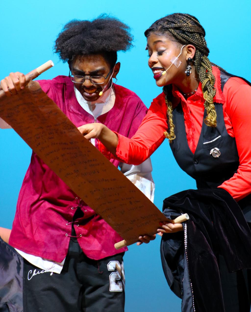 Two performers on stage, a young person and a woman, examine a long scroll.