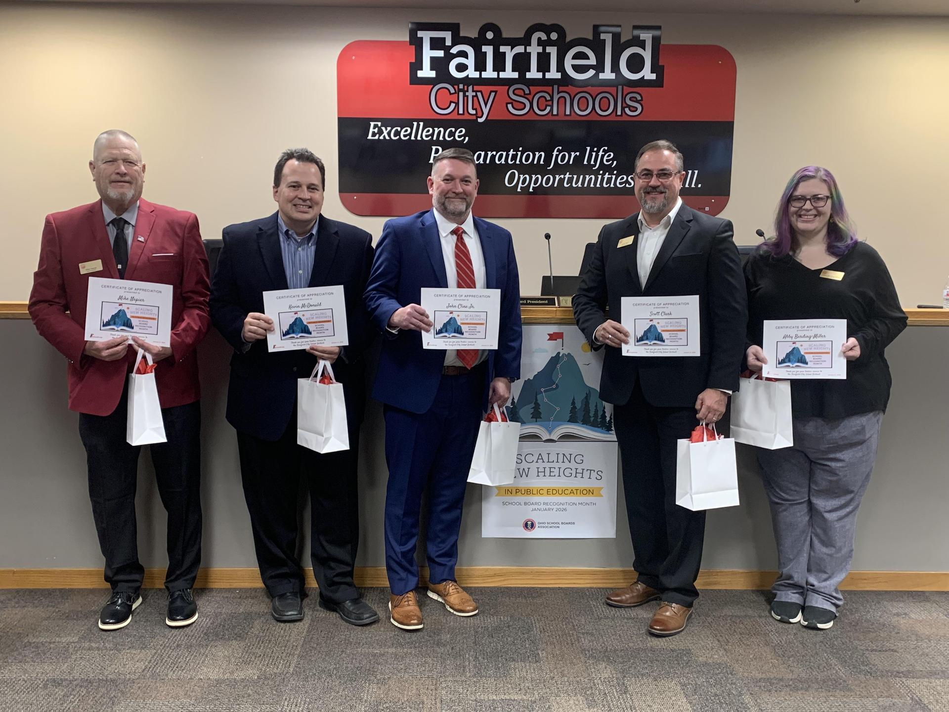 Board members stand in front of a 'Fairfield City Schools' sign, each holding a certificate and a white bag.