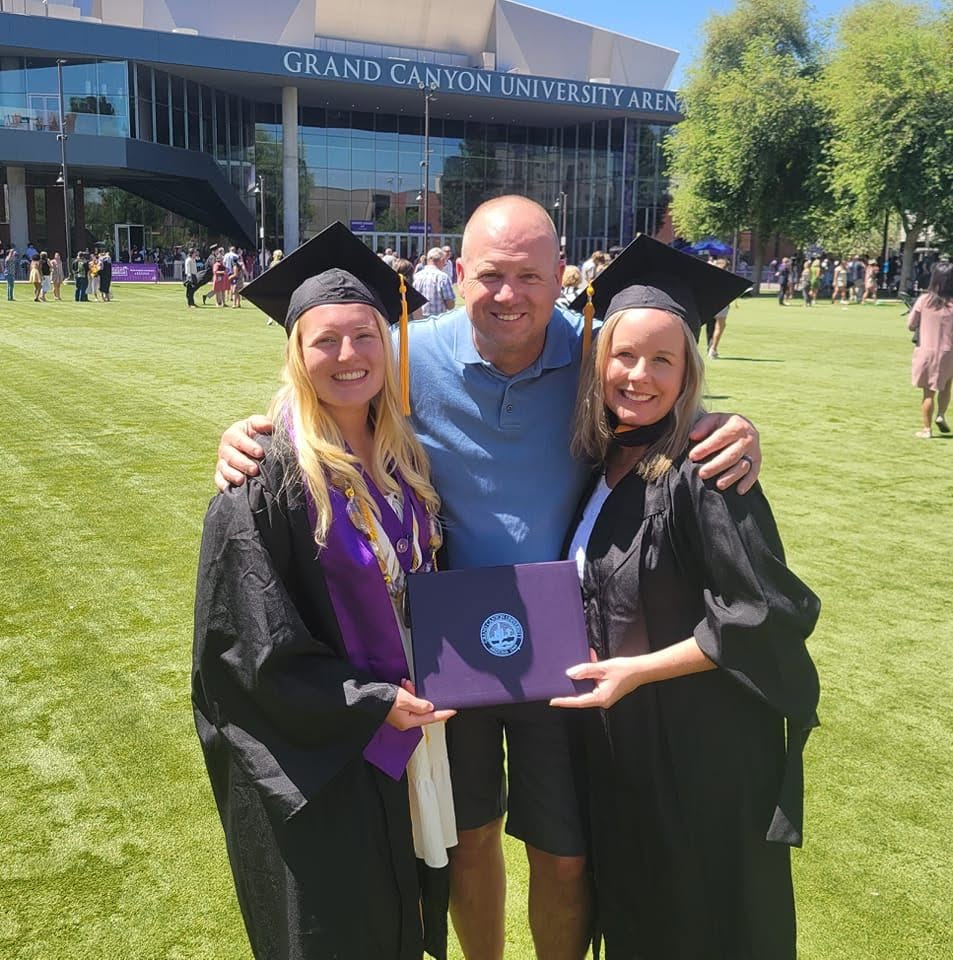 Terry poses with his two daughters in graduation gowns and caps on a sunny day.