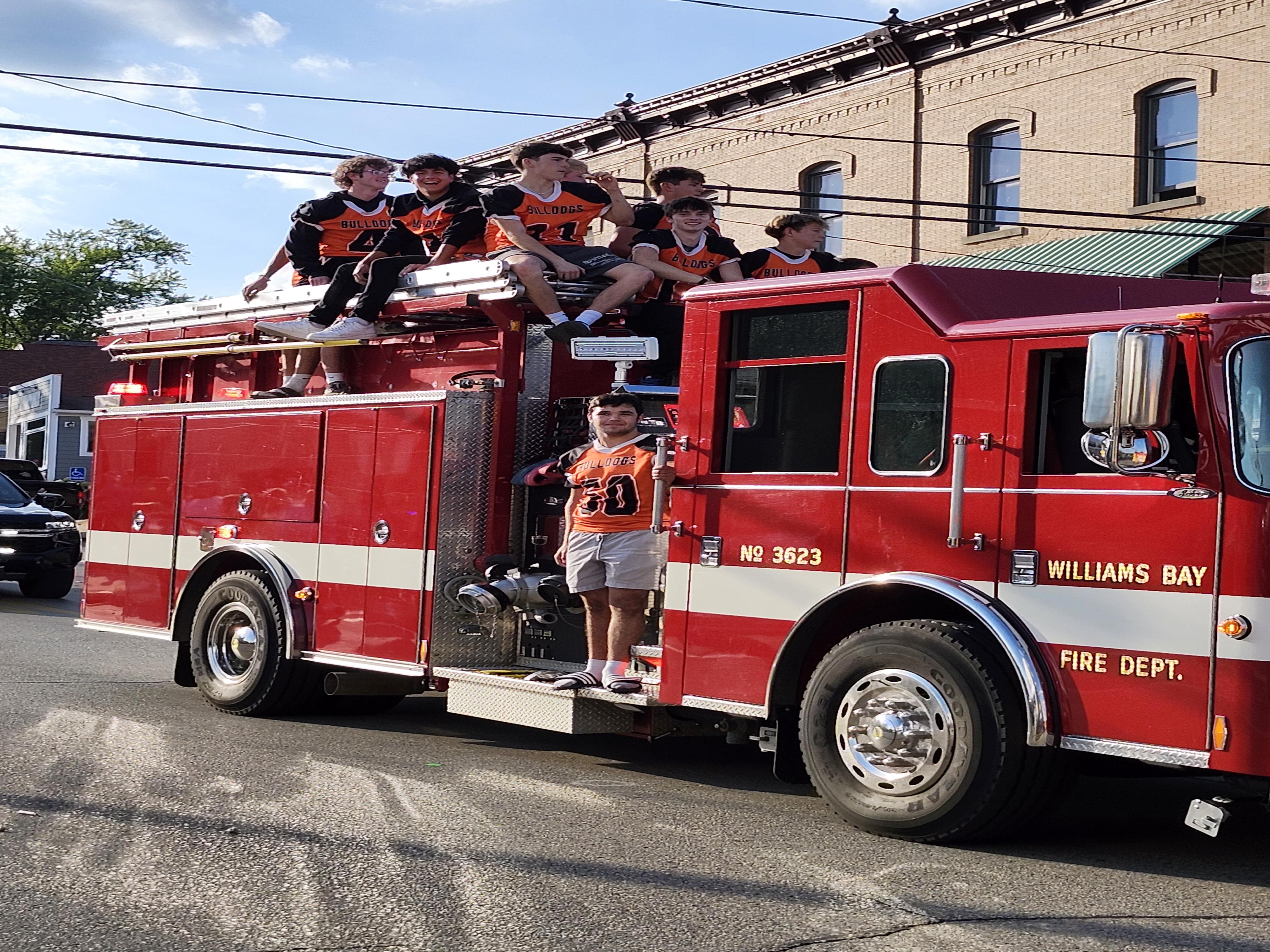 A bright red fire truck with several people riding on top, participating in a parade.