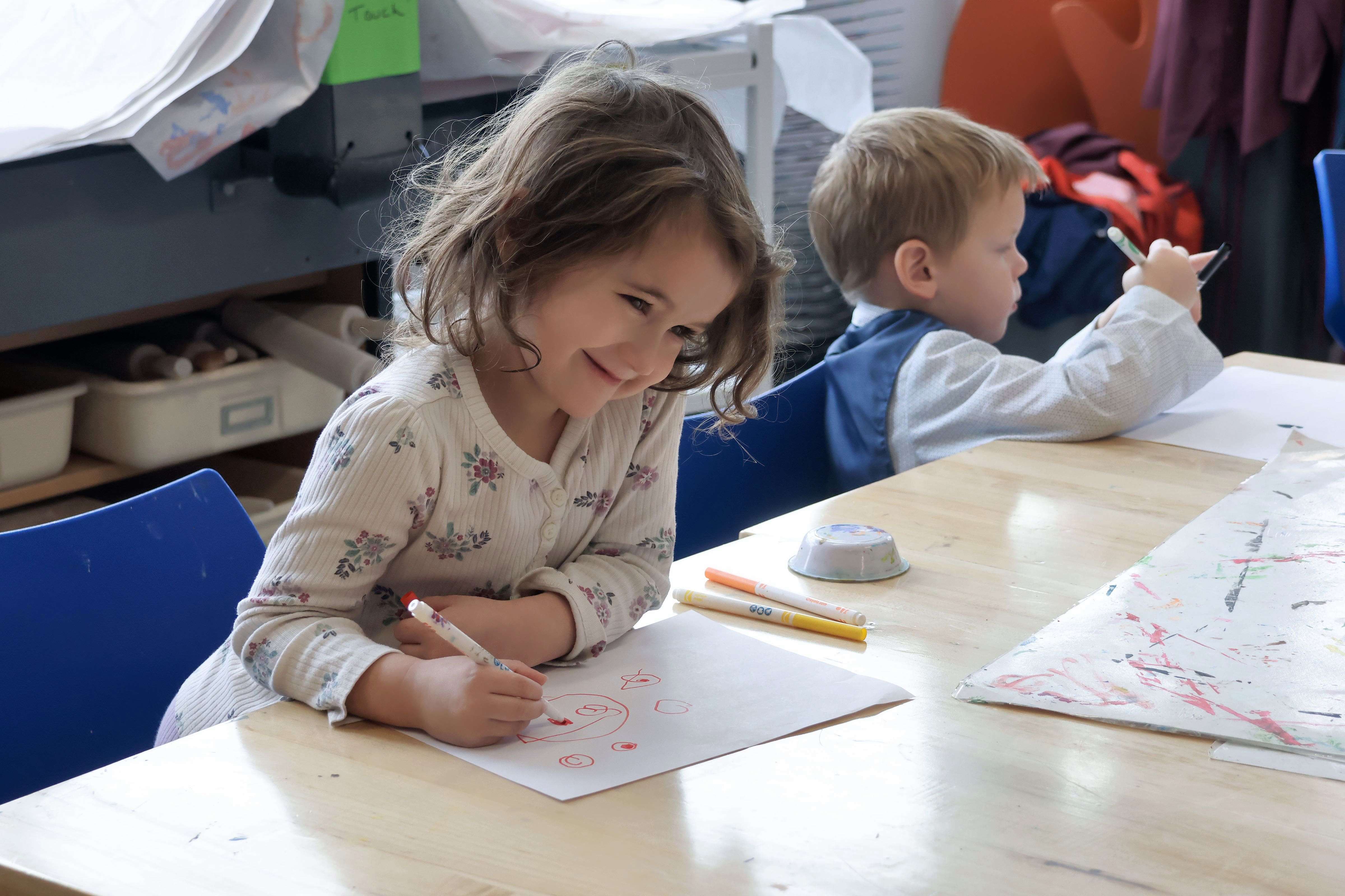 A young girl with brown hair smiles as she draws on a piece of paper at a table.