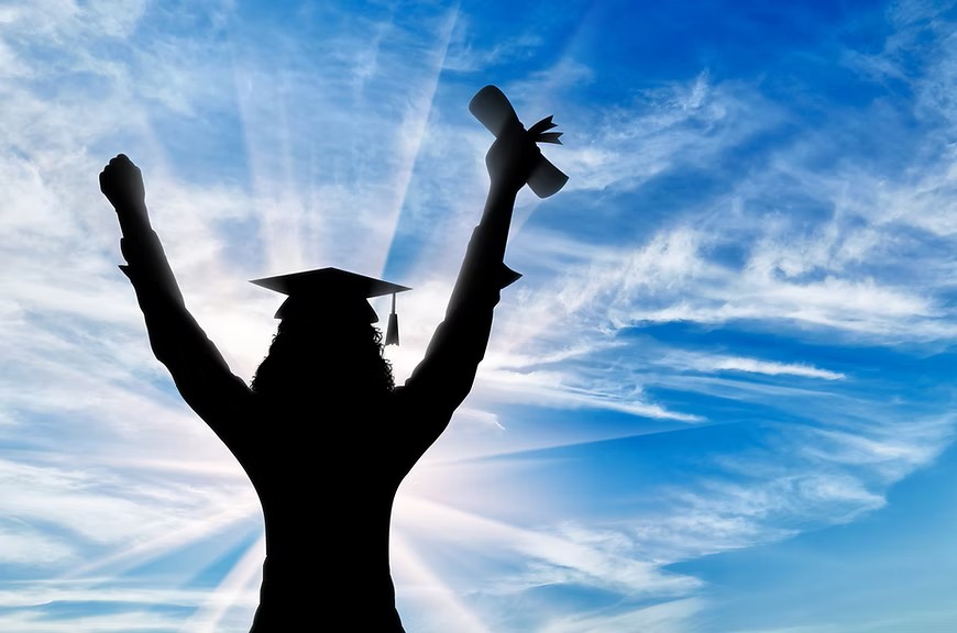 Silhouette of a graduate in a cap and gown raising a diploma in the air against a bright, cloudy sky.