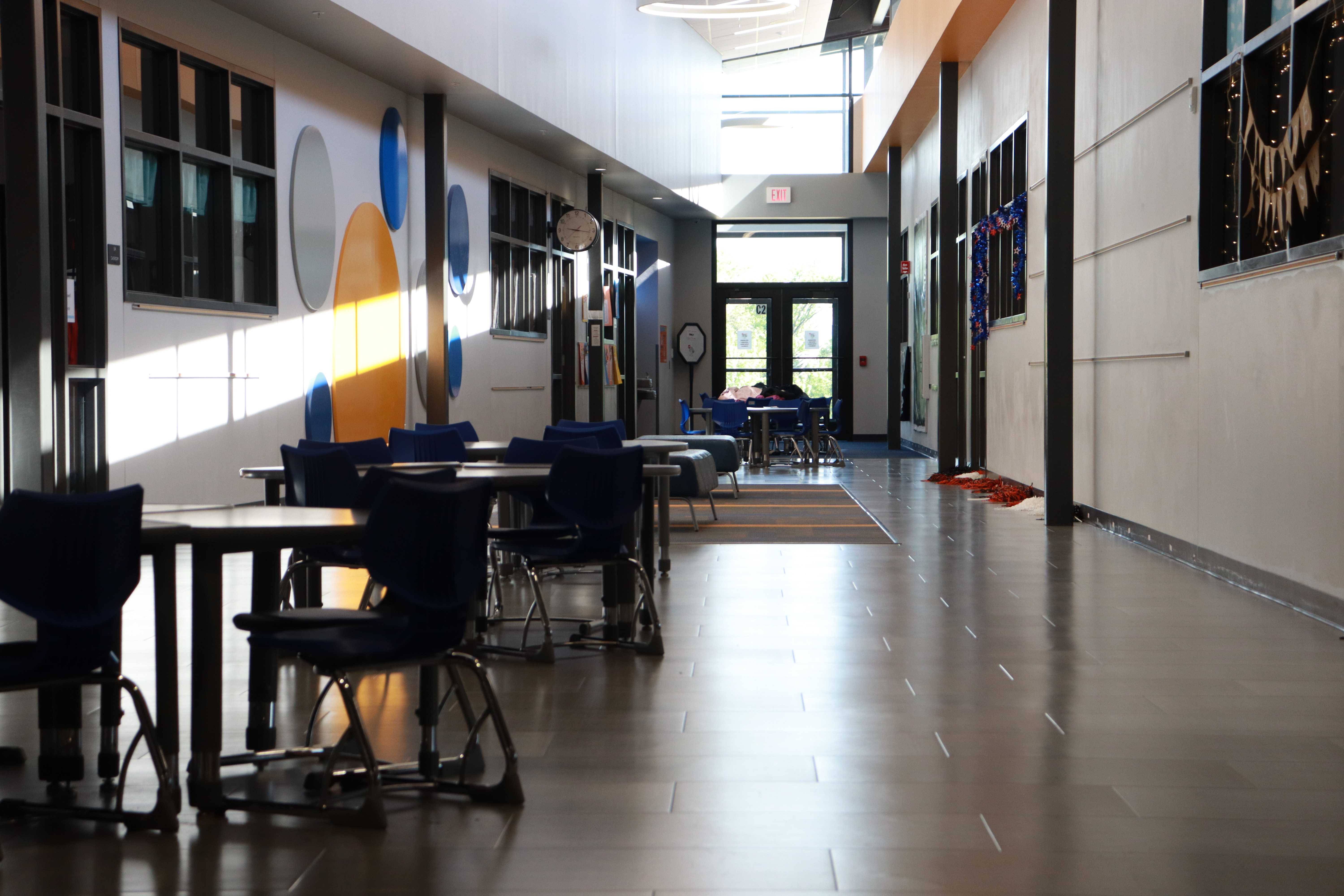 A modern school hallway with tables and chairs, bathed in sunlight.
