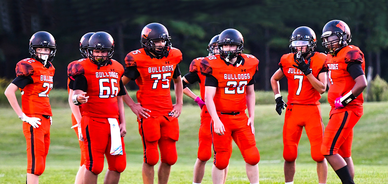 A group of young football players in orange uniforms stand on a grassy field.