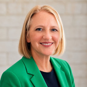 A woman with blonde hair smiles at the camera, wearing a green blazer.