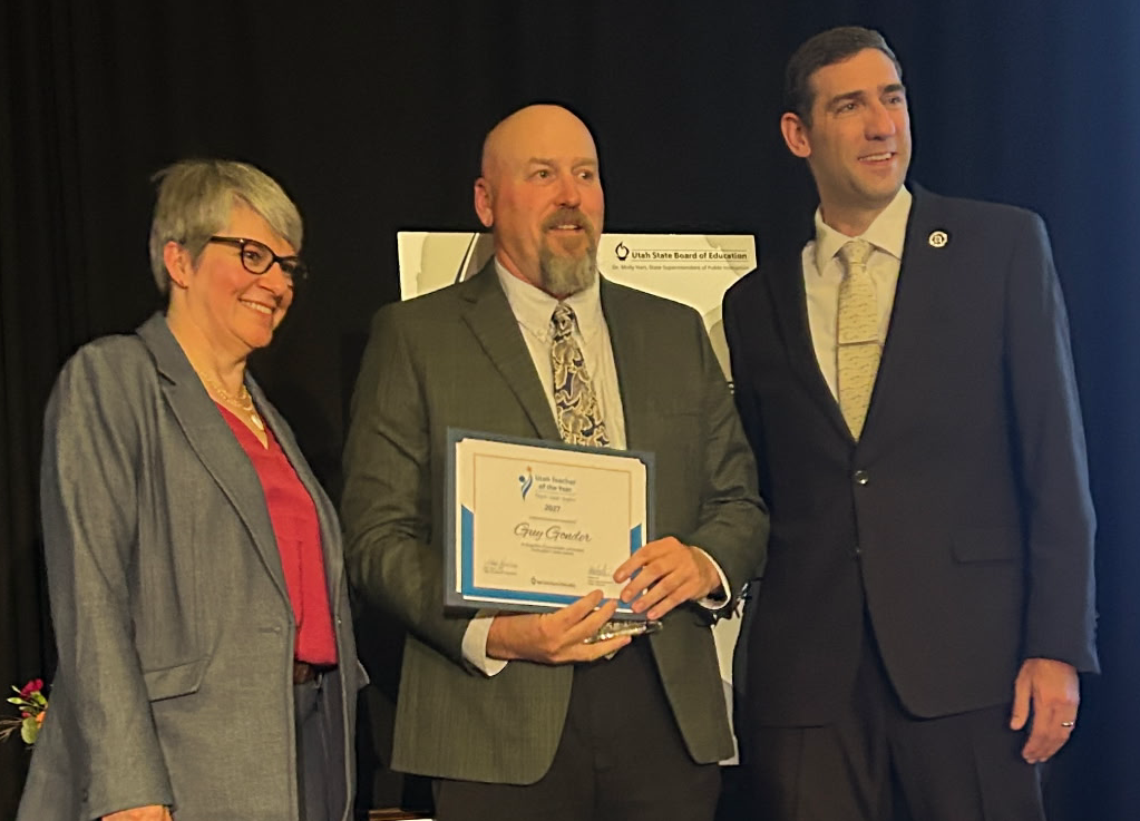 A man holding a certificate stands proudly between two colleagues on a stage.