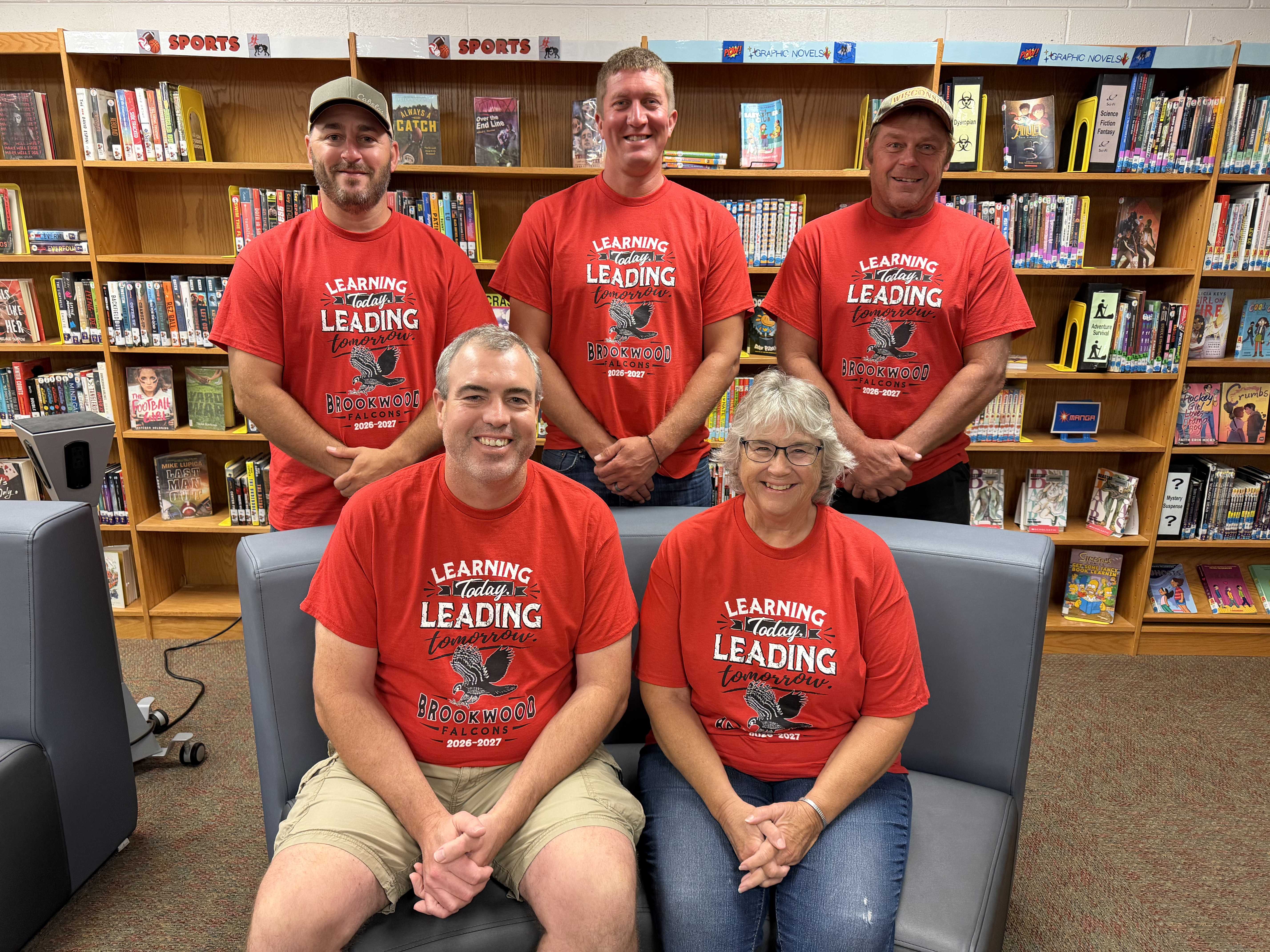 Five people pose together inside a library, all wearing matching red t-shirts with text.
