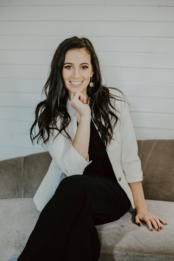 Dr. Leadbetter smiles confidently while sitting on a couch, wearing a white blazer over a black outfit