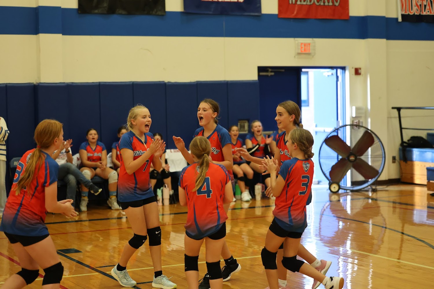 A group of young female volleyball players in red and blue uniforms celebrate on a court.