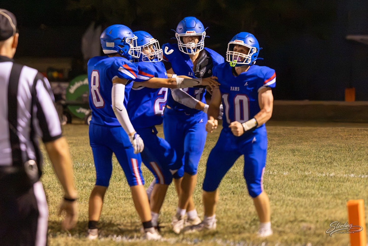 A group of football players in blue uniforms celebrate on the field during a night game.