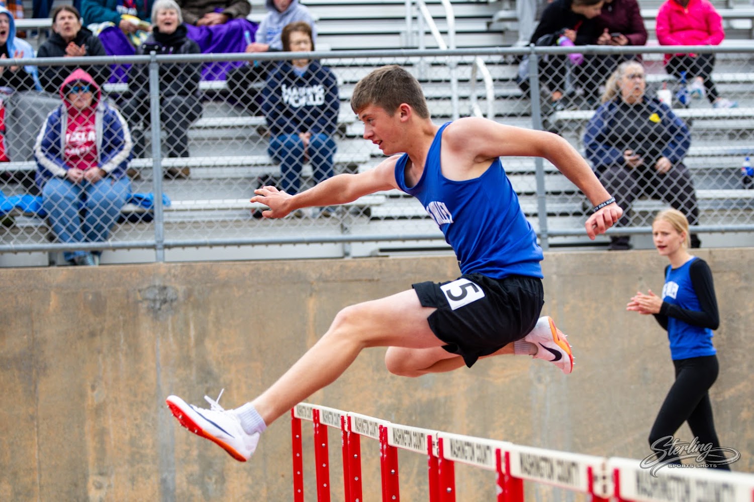 A male runner in a blue singlet leaps over a hurdle during a track and field race.
