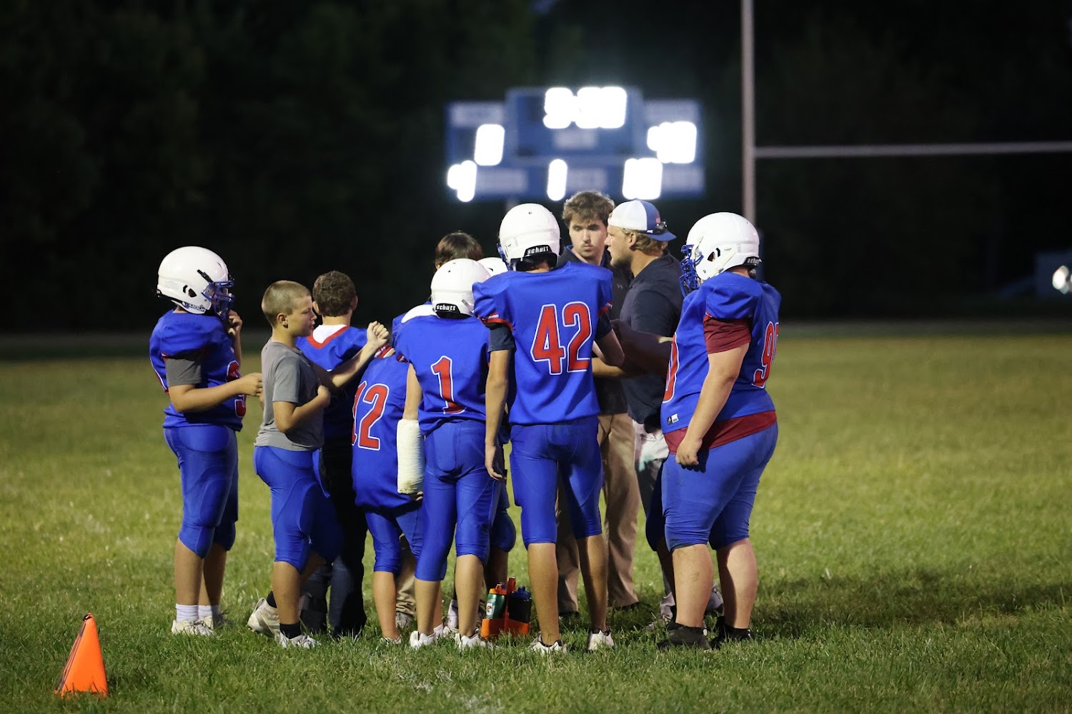 A football team huddles together on a field at night, with a scoreboard visible in the background.