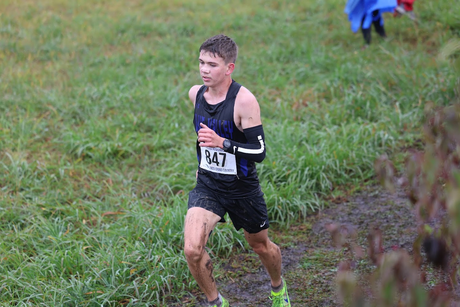 A young male runner in a black singlet and shorts competes in a cross-country race.