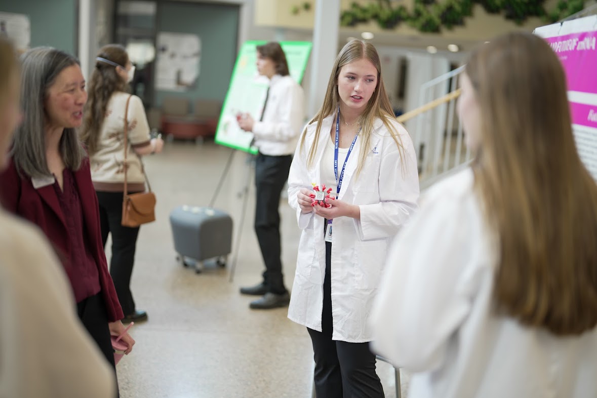 Students in white lab coats converse in a brightly lit hallway during a poster presentation event.