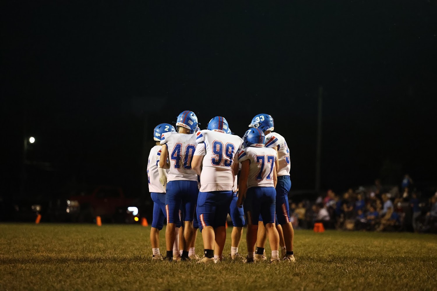 A football team huddles together on a field at night, wearing blue and white uniforms.