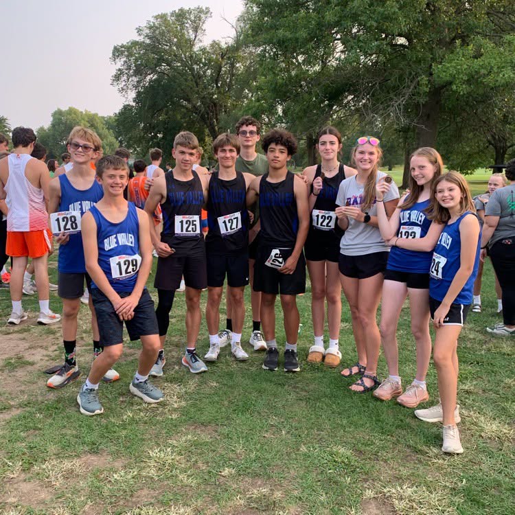 A group of young athletes in matching blue uniforms stand together outdoors, some wearing race bibs.