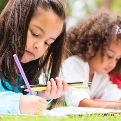 Two young girls lie on the grass, focused on drawing with colored pencils.