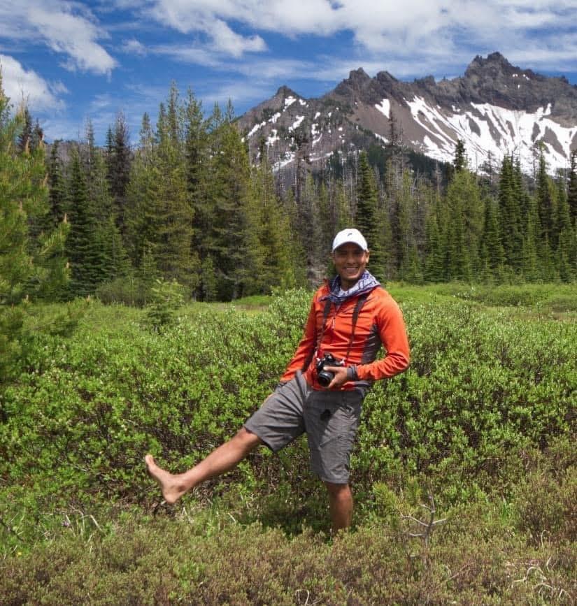 Carlos Sequeira on a hike in a field in front of mountains, holding a camera