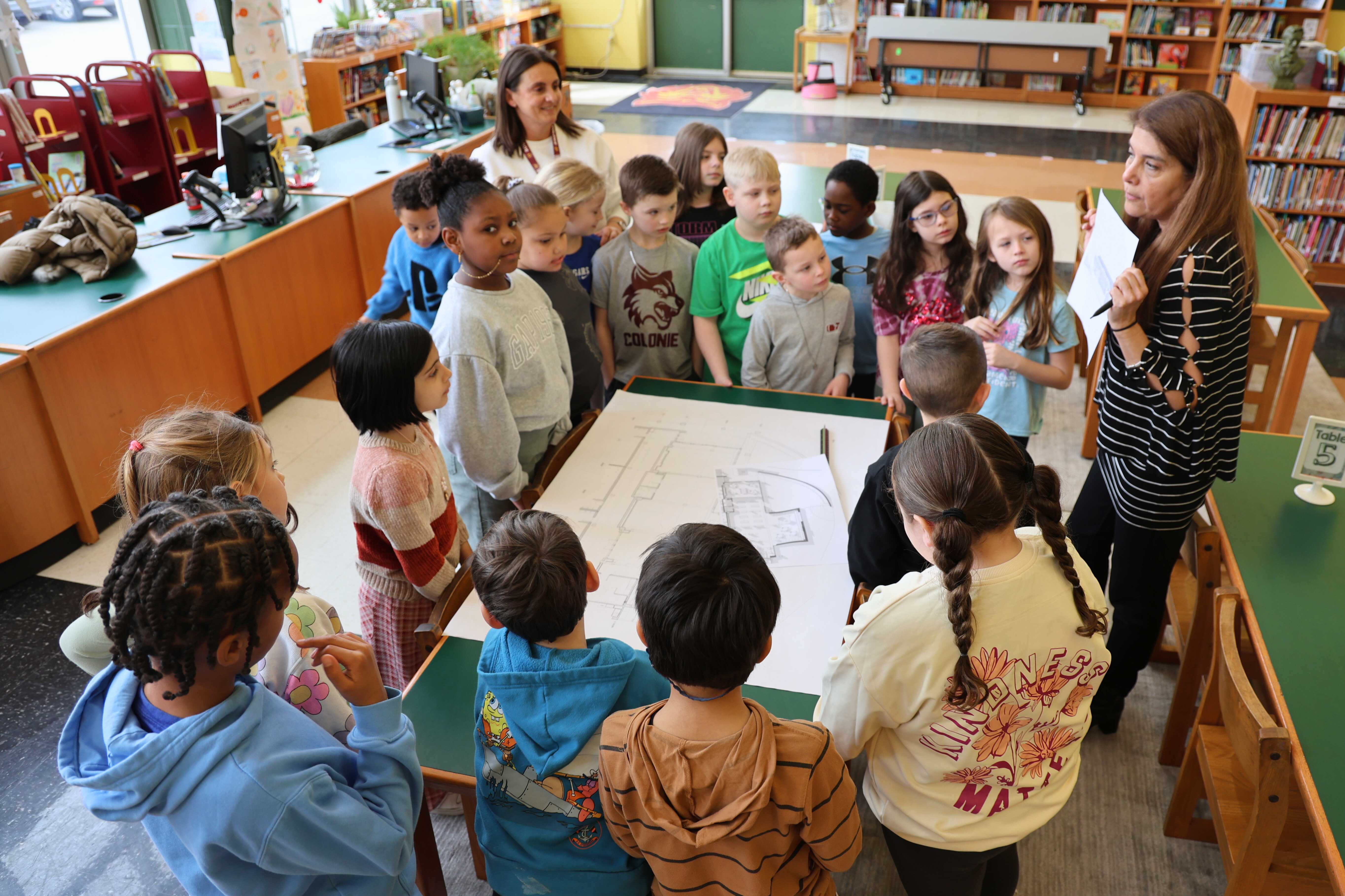A group of children gather around a table, listening to an architect holding up a plan.