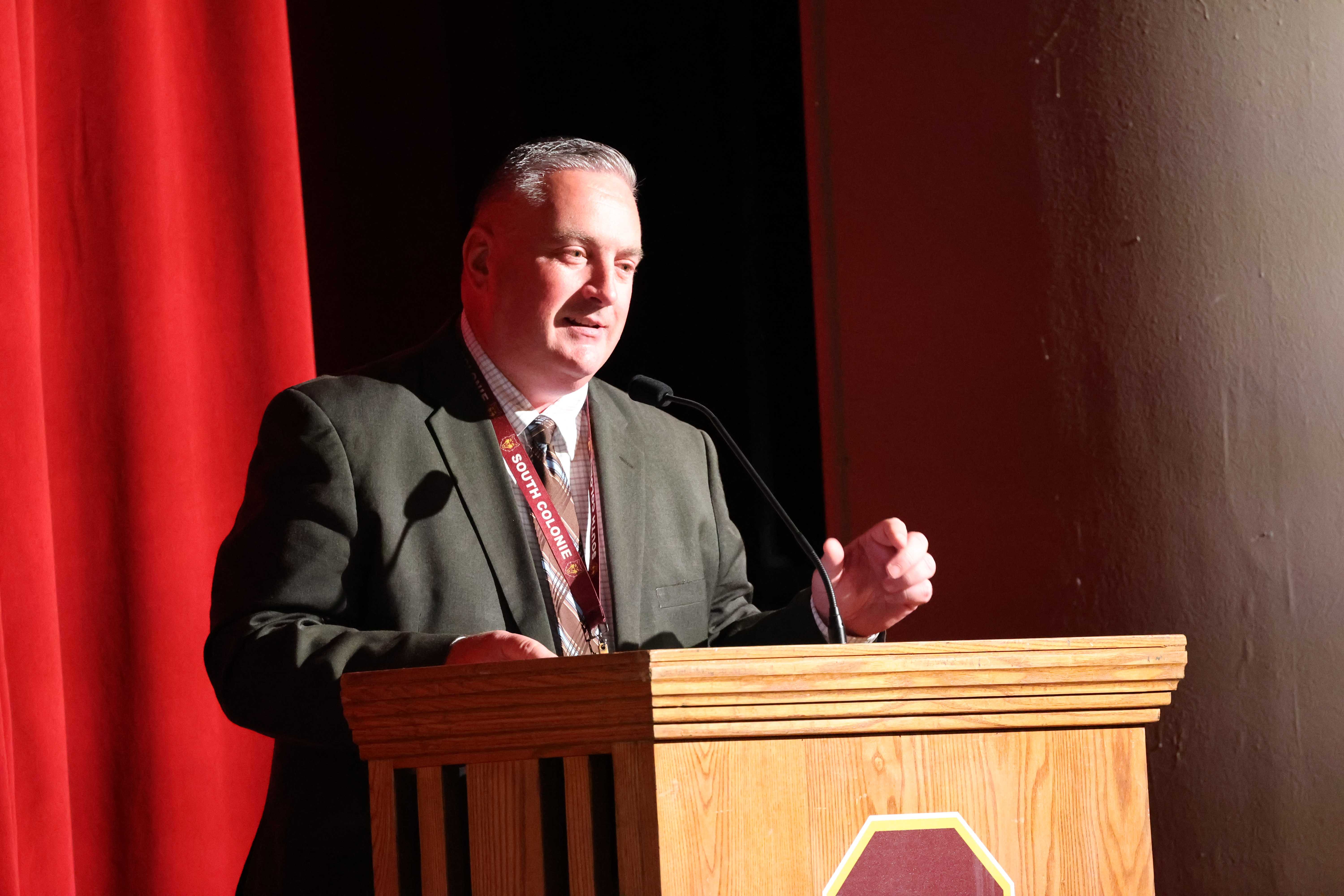 A man in a suit speaks at a wooden podium on a stage with a red curtain.