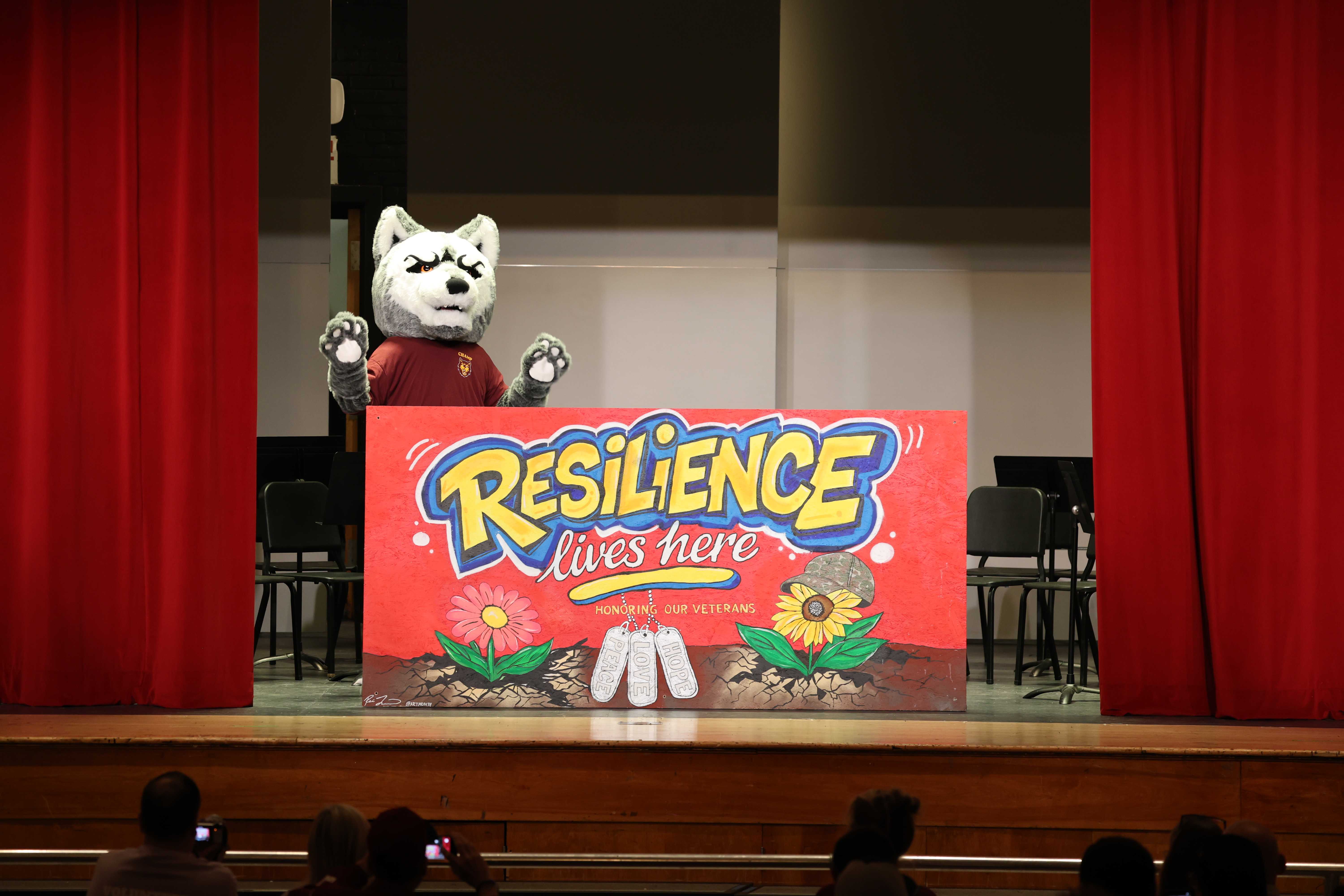 A school mascot stands behind a sign that reads 'Resilience lives here' on a stage.