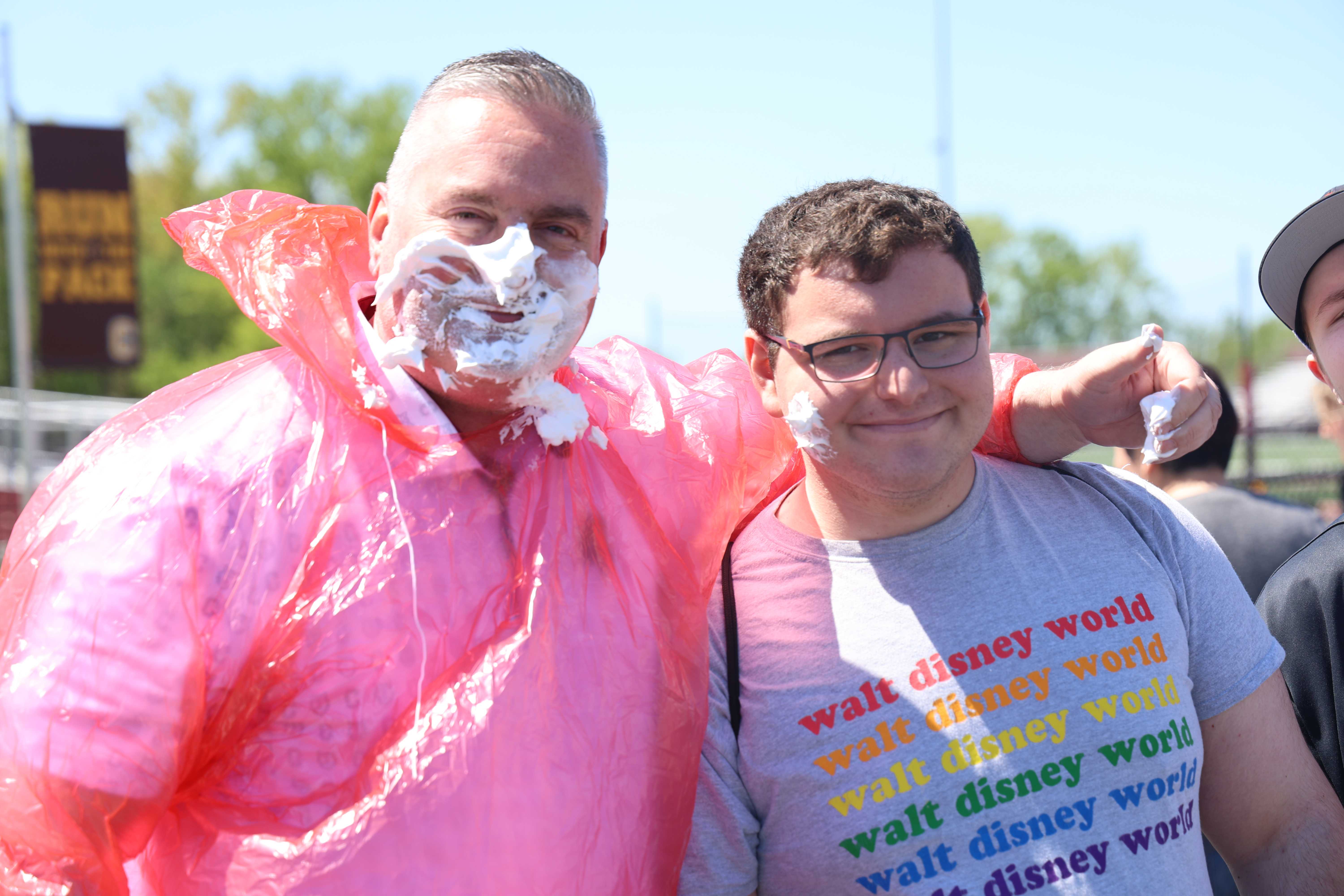 Two men smile, both with shaving cream on their faces, one wearing a pink poncho.