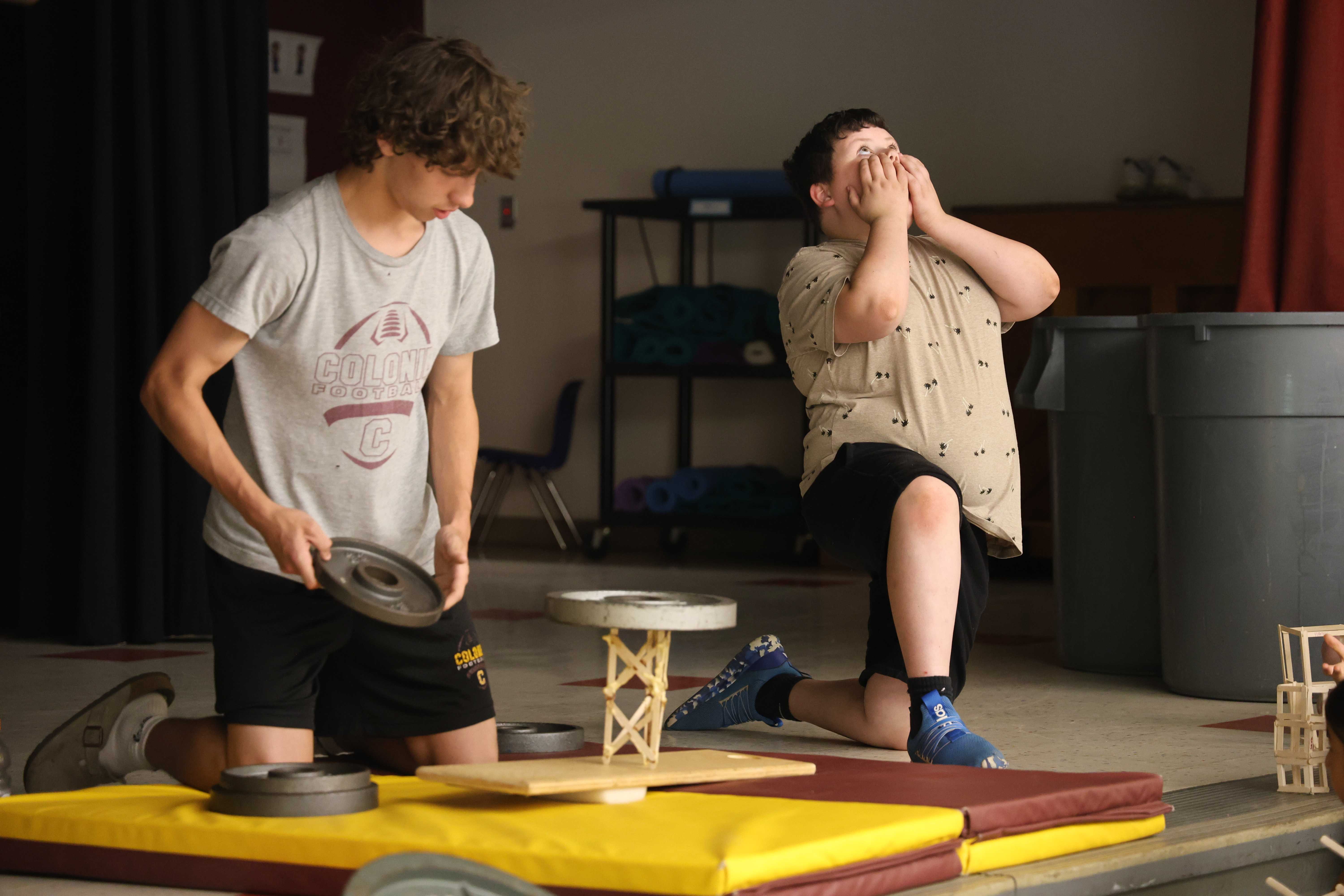 Two boys are kneeling on mats, one holding a weight plate and the other with hands over his mouth in surprise.