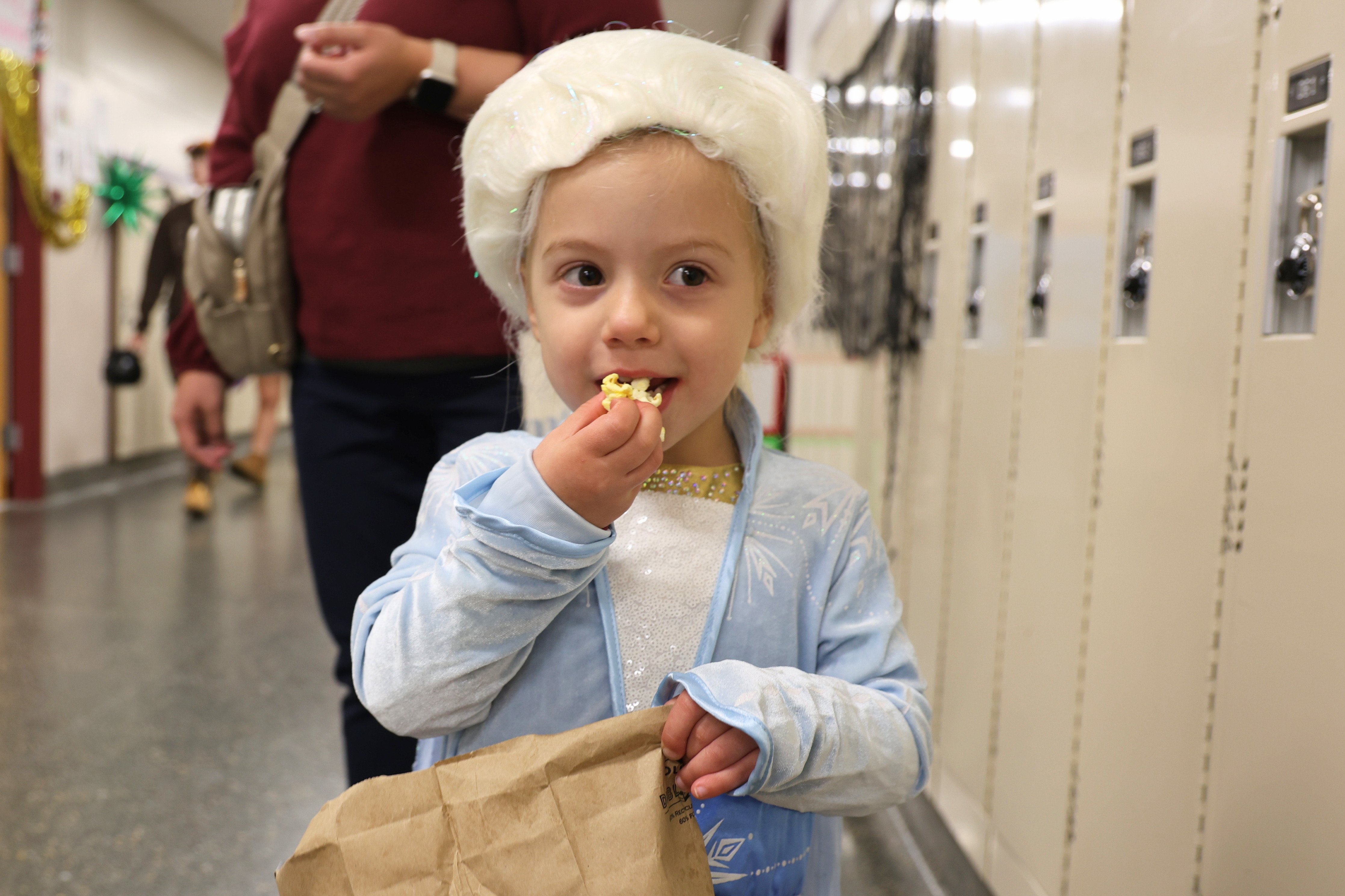 A young child wearing a white wig and Elsa costume eats a snack from a brown paper bag in a hallway with lockers.