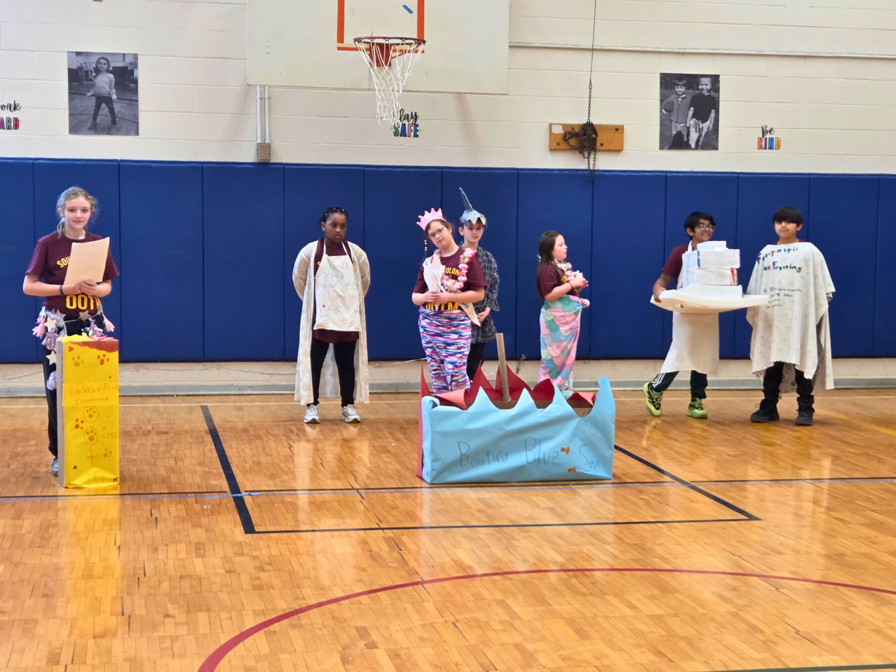 A group of students stand on a basketball court, some holding props.