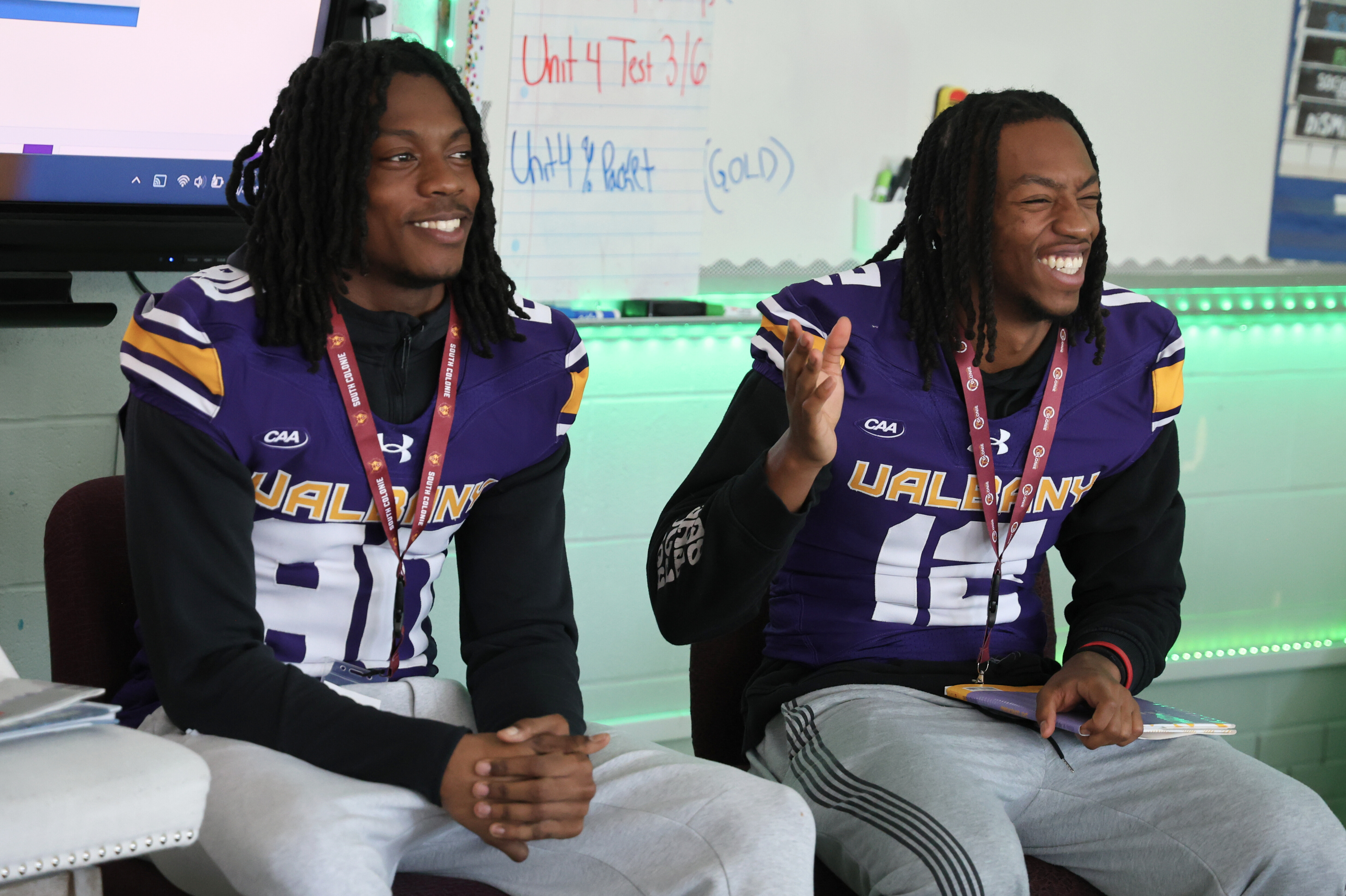 Two football players in purple and gold jerseys smile and laugh in a classroom