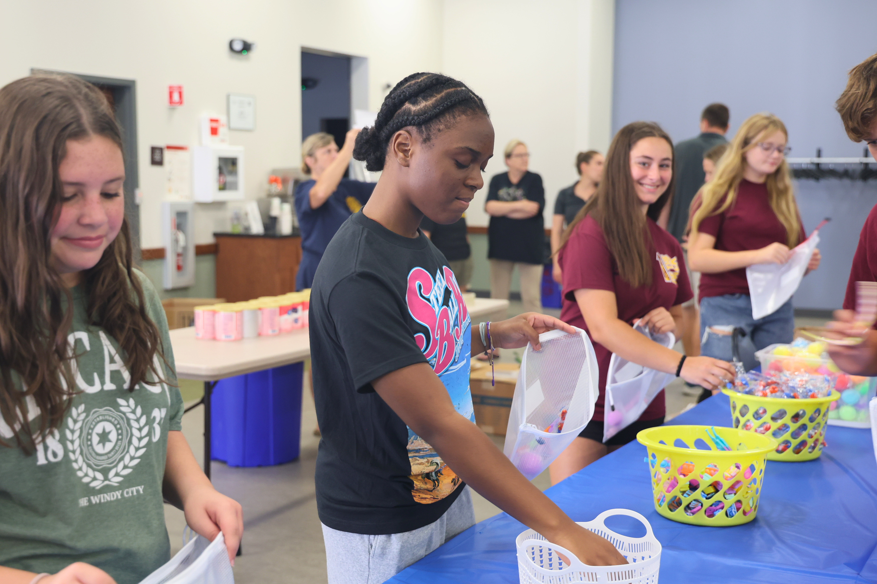 A group of students pack items into containers at tables in a brightly lit room.