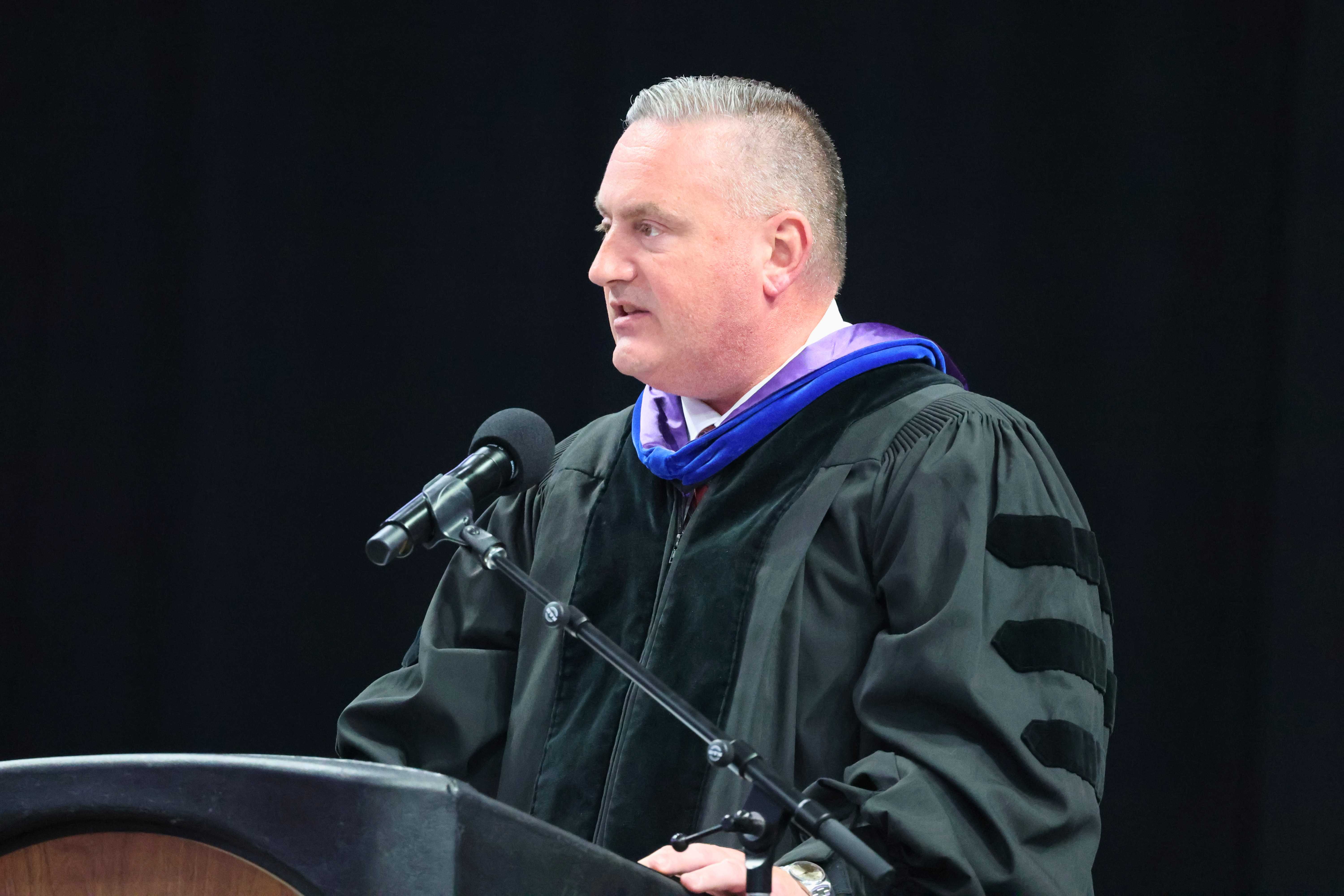 A man in academic regalia speaks at a podium with a microphone.