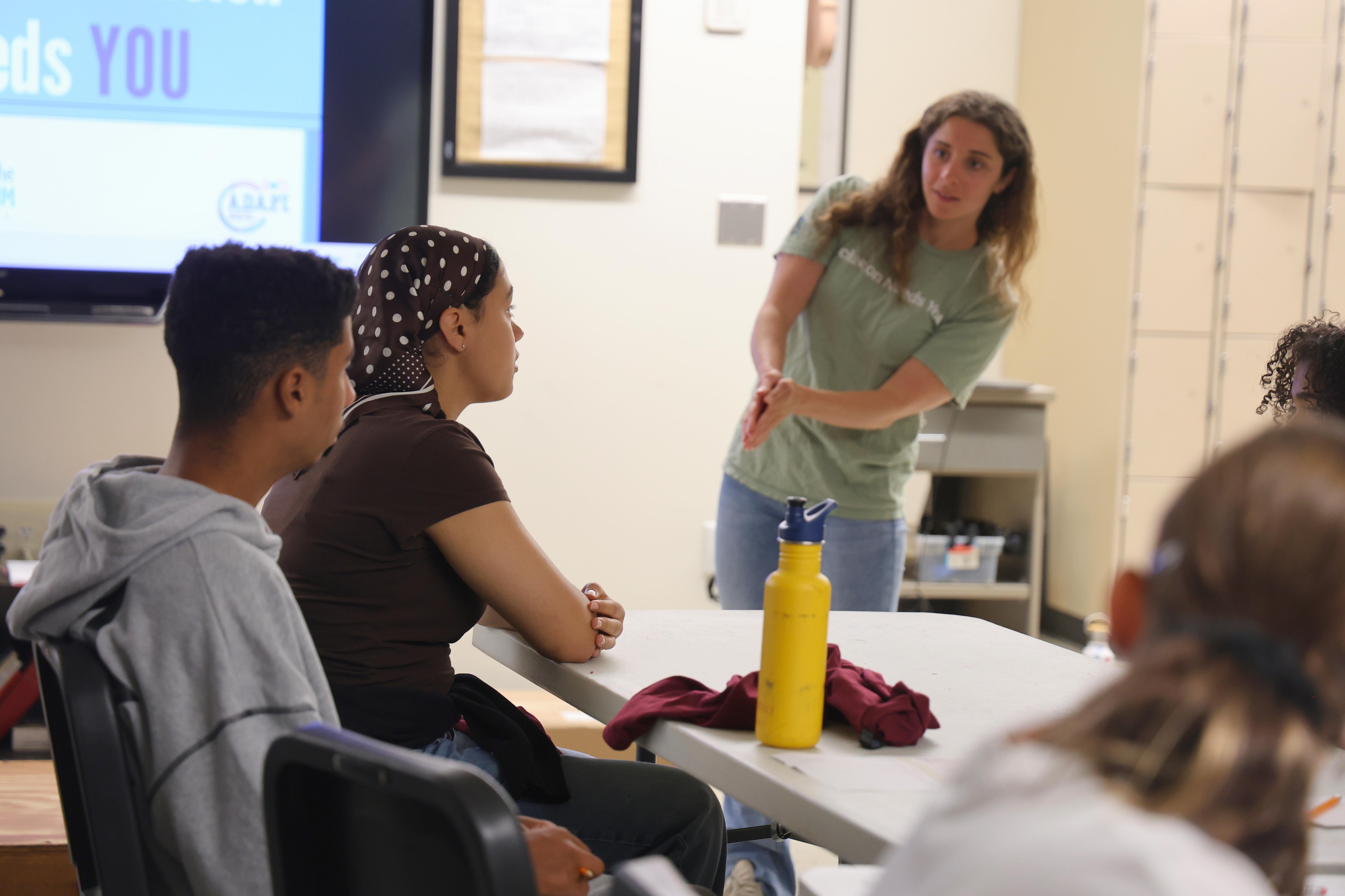 A group of people sit around a table in a bright room as one person stands to present.